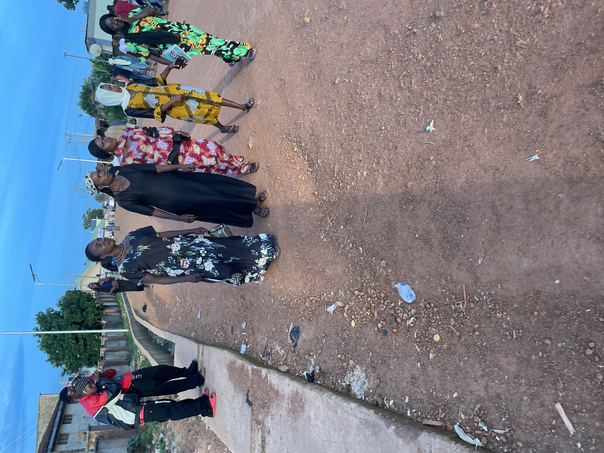 The image captures a street scene in Gura Top, Nigeria, under a twilight sky. A group of individuals, predominantly women dressed in vibrant, patterned attire, are walking on a dirt road. The women are adorned in long dresses and head coverings, with one woman in a yellow and grey patterned dress holding a book or pamphlet. To the left of the frame, two young men are standing on a concrete curb. One man, wearing a red and black jacket with white accents, has his arm around the other who is wearing a red shirt and a patterned beanie. The road is unpaved and dusty, with scattered debris and small rocks. In the background, buildings and streetlights are visible, suggesting an urban or semi-urban setting. The lighting indicates it is either early morning or late evening.