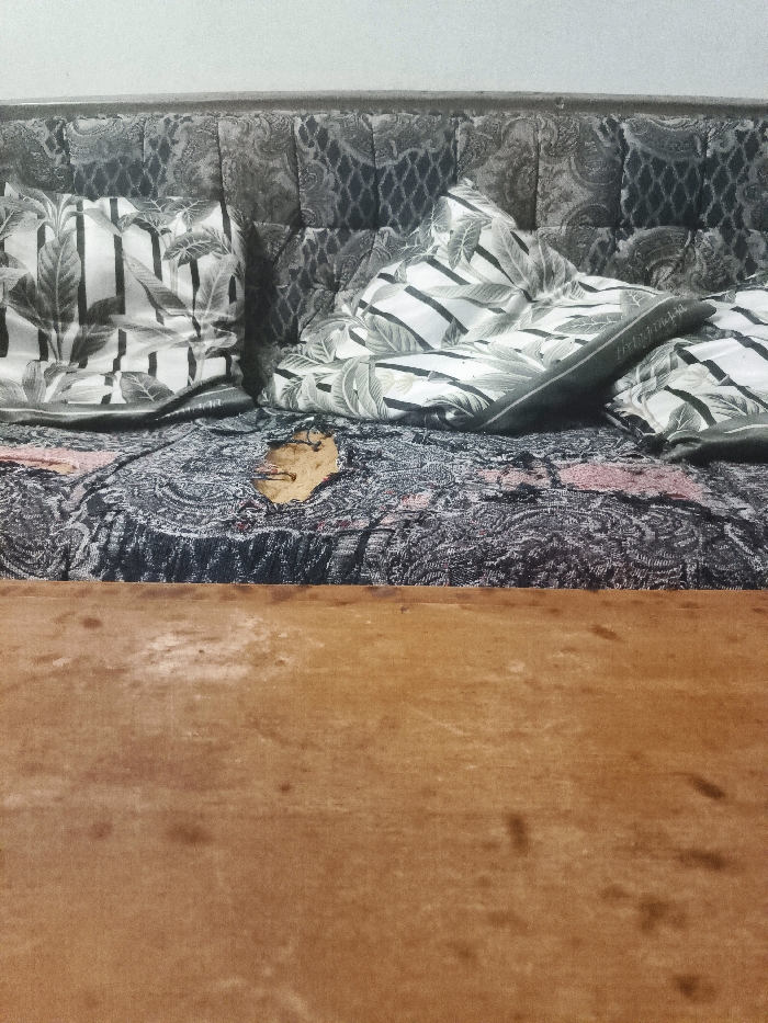 An indoor scene in Cimenyan, Indonesia, displays a sofa or daybed positioned against a plain, light-colored wall. The sofa features a dark, patterned upholstered backrest. Its seat is covered by a dark blue and black patterned blanket or throw with subtle red or pink accents. A significant, irregular tear in this blanket, approximately 15-20 cm in its longest dimension, exposes a lighter brown material underneath. Two pillows with black and white leaf patterns are arranged on the sofa; one on the left features vertical stripes, and the one on the right is partially obscured by a folded, dark green or grey fabric item. The lower half of the image reveals a light brown wooden surface, possibly a table or the front frame of the furniture, exhibiting wear and faint marks. No individuals are present, and no actions are in progress.