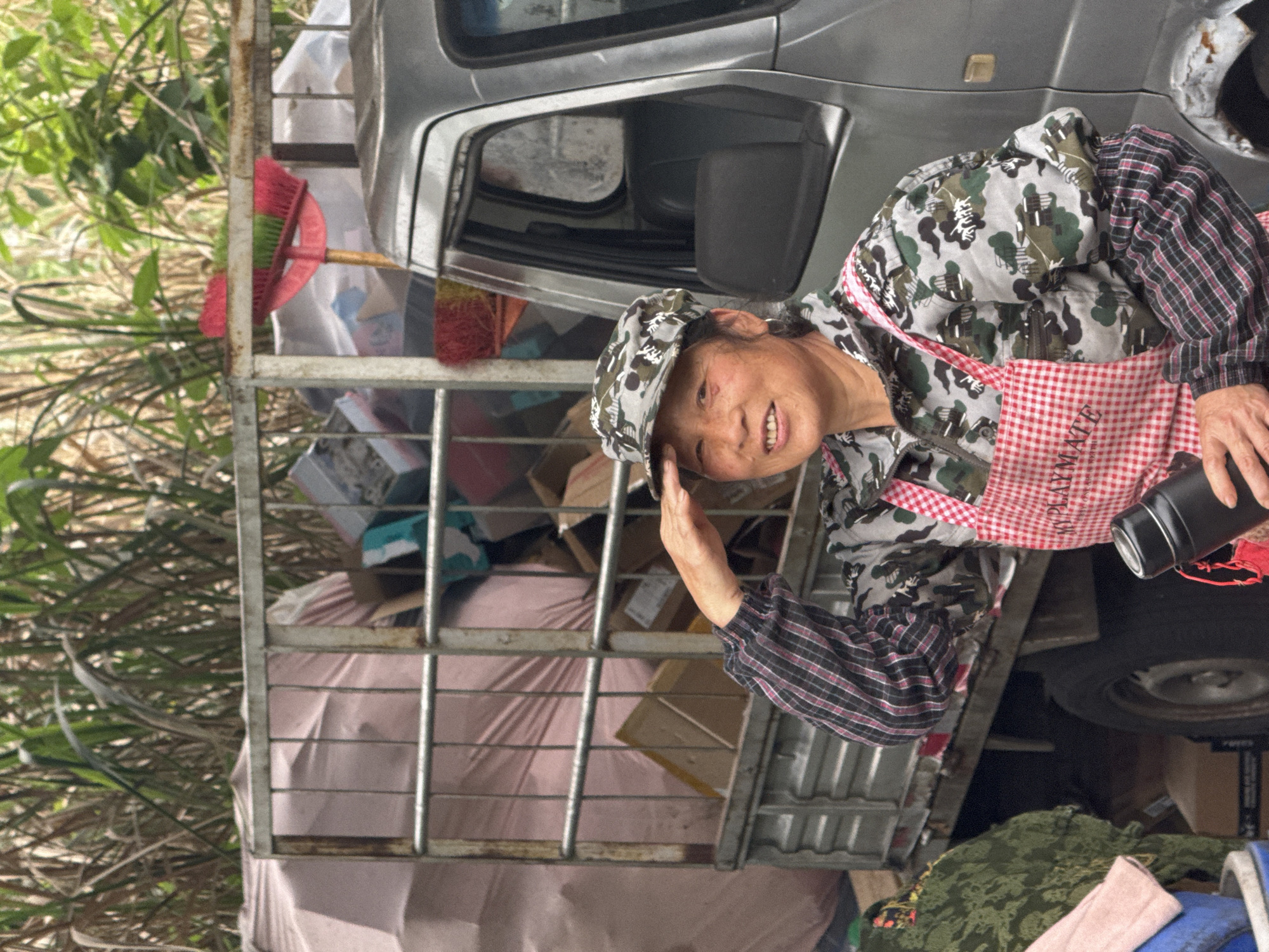 A woman with a camo cap and camo jacket with plaid sleeves, and a red and white checkered apron stands next to the passenger side of a silver utility truck. The truck is full of various goods secured by a metal cage structure. The woman is smiling, holding her hand to her head in a partial salute while holding a black thermos in the other. Green vegetation is visible in the background. The apron says "PLAYMATE". The scene is located in Jiangjin, China.