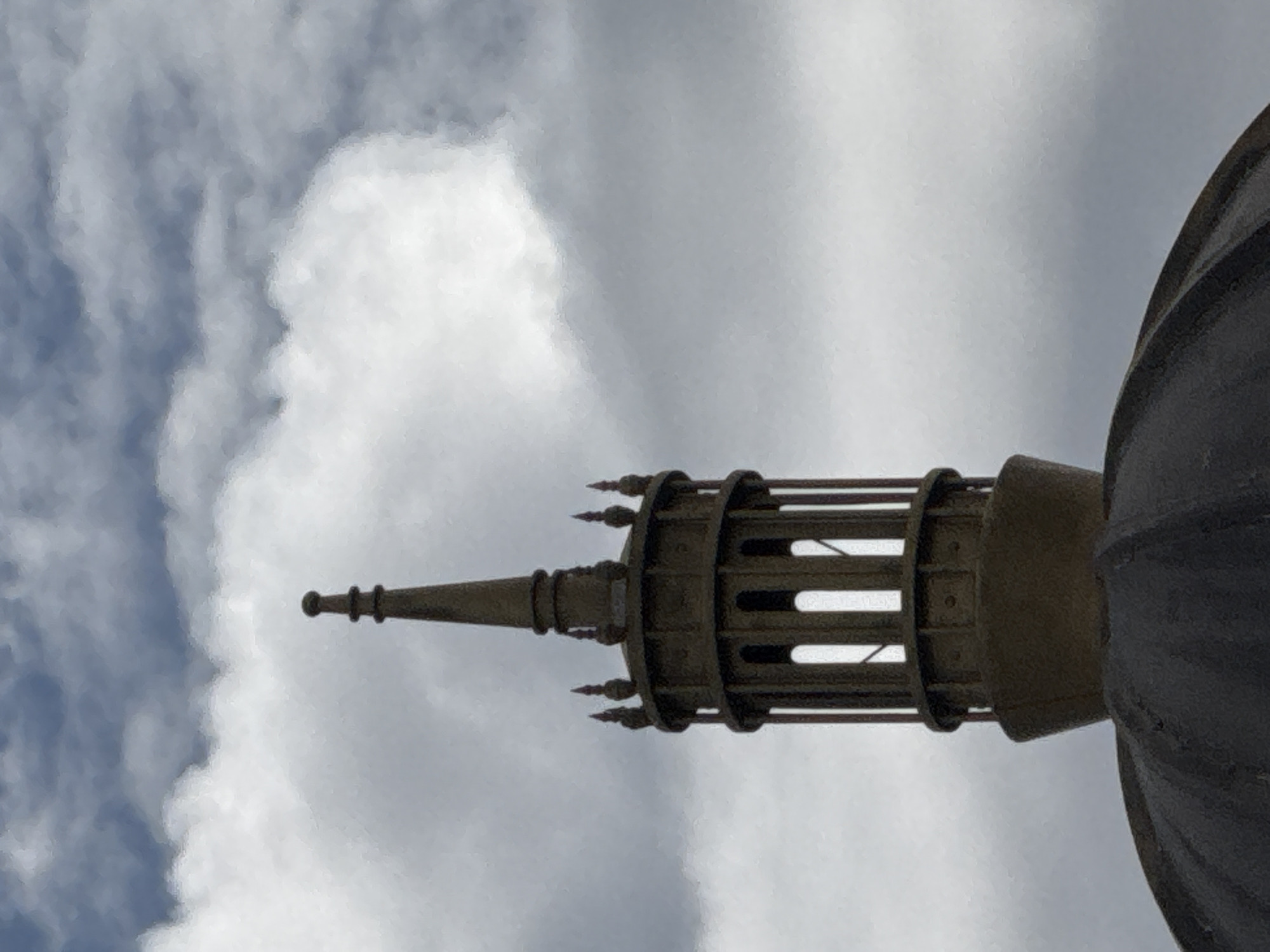 The image shows a close-up of a dome topped by a decorative turret against a cloudy sky. The turret features vertical slats and a finial at its apex. The dome itself is dark and appears to be made of metal. The overall scene is dominated by the architecture and the sky. The location is identified as Da Lat, Vietnam. There are no people, actions, or interactions depicted in the image.