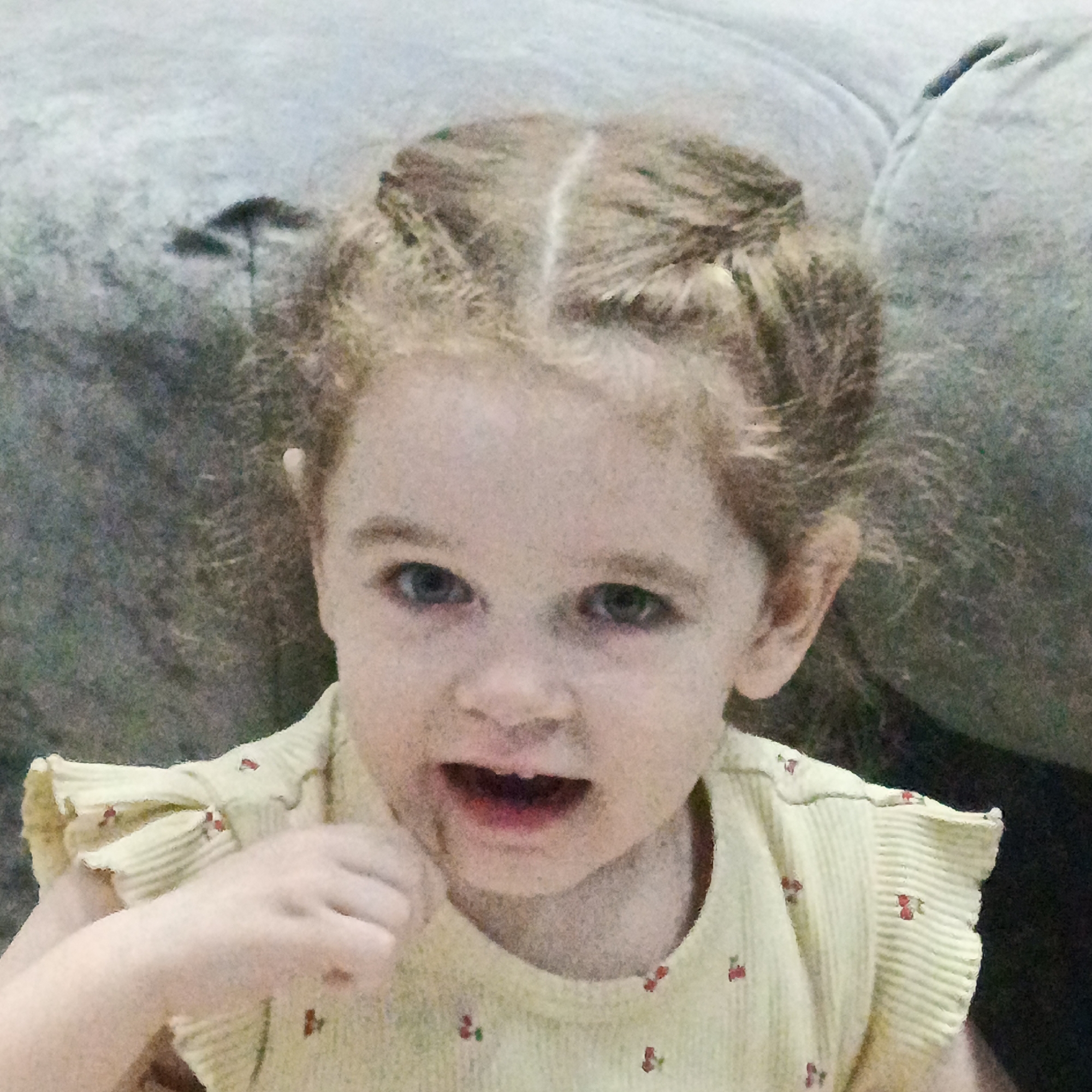 A young child, appearing to be a toddler, is the primary subject of the image. She has blonde hair styled in two buns on top of her head. The child is wearing a yellow shirt with a small cherry pattern. She is holding something small near her mouth, possibly a small object. Her expression is neutral to slightly surprised.
The background consists of a blurred, light-colored couch or upholstered furniture. The image appears to be taken indoors.
The location of the image is not evident from the image itself. There are no distinctive features within the image to confirm it was taken in the city of Марковский, Russia. The location detail cannot be confirmed.
