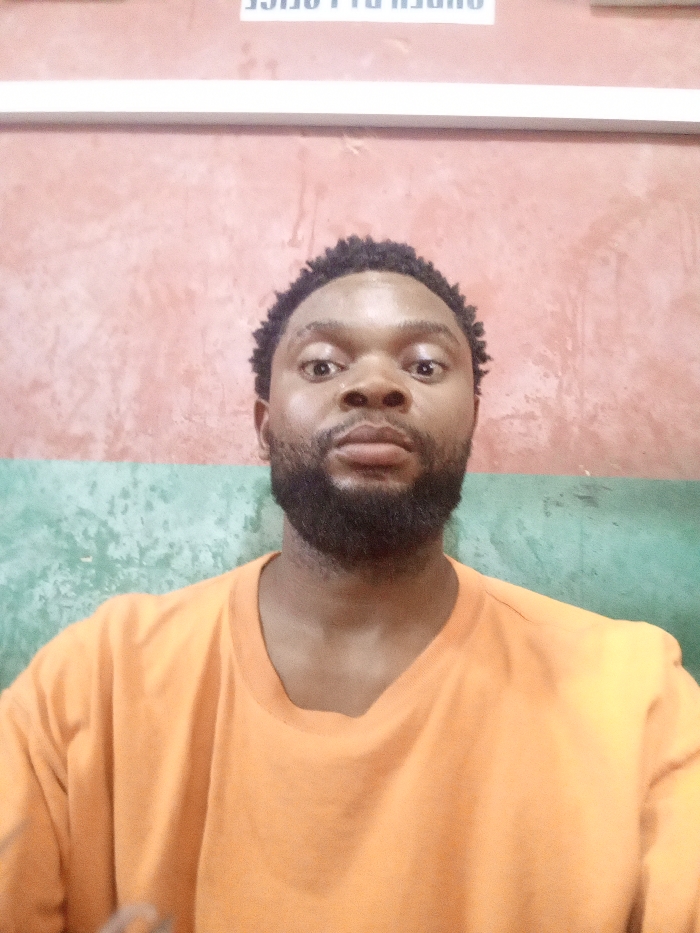 The scene is an indoor location in Magboro, Nigeria. An adult male is centered in a medium close-up, facing the camera directly. The individual has dark skin, short dark curly hair, and a full, dark beard. He is dressed in a plain orange crew-neck t-shirt. His eyes are wide-set and open, maintaining direct eye contact with the viewer; his mouth is closed in a neutral expression. The background is a painted wall, horizontally segmented. The upper portion is a faded reddish-pink, and the lower portion is a faded teal green. A white horizontal trim piece is affixed above the subject's head, and inverted white lettering, which reads 'OFFICE' in reverse, is partially visible above this trim.