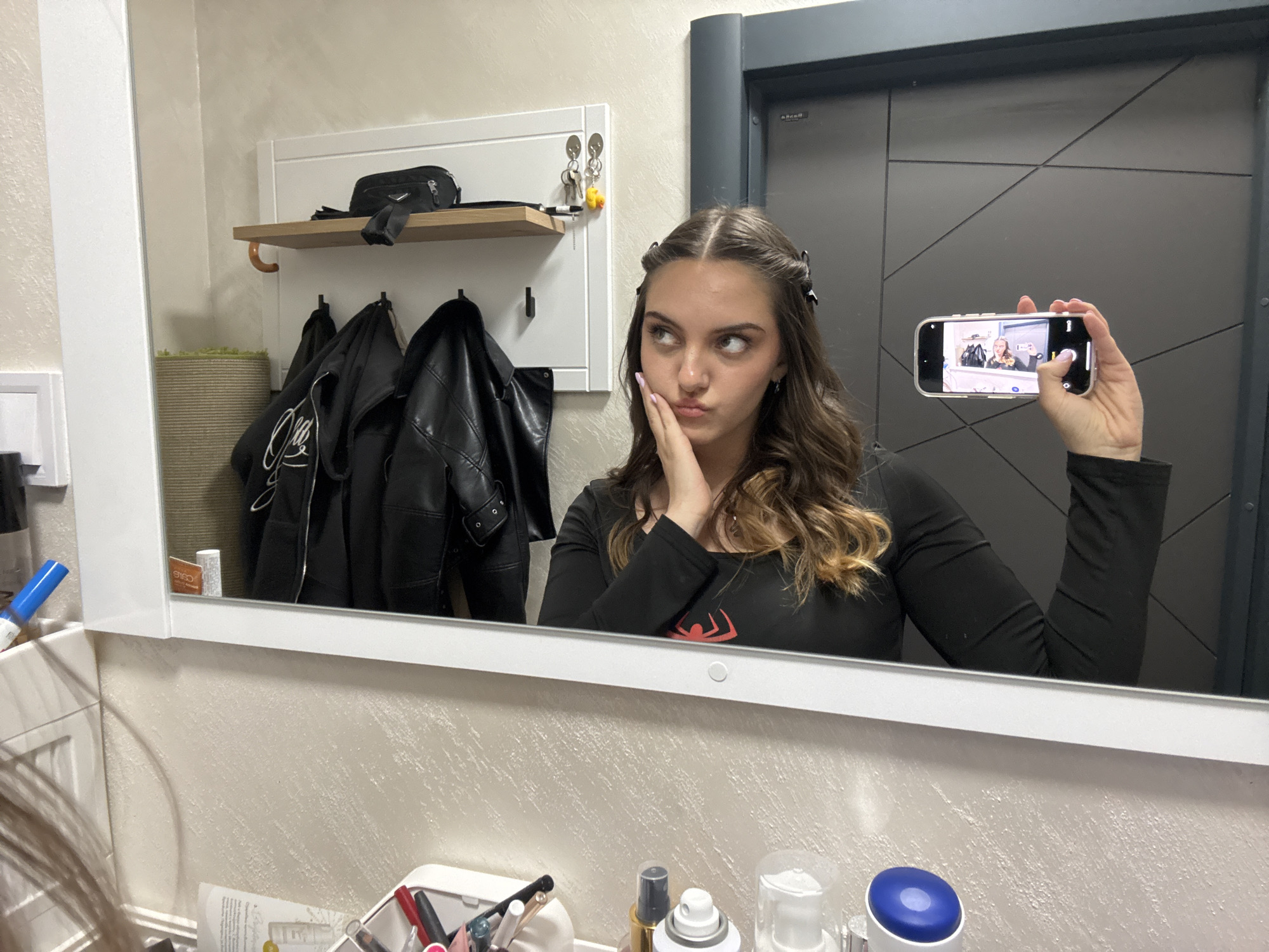 A young woman is taking a mirror selfie. She is holding a smartphone in her right hand, capturing her reflection in a bathroom mirror. She is wearing a black long-sleeved shirt with a red Spider-Man logo. Her hair is brown with blonde highlights, and she has clips on both sides of her head. Her left hand is resting on her cheek, and she has a contemplative expression. Behind her, there's a white wall rack with a shelf holding a small black bag. Several black jackets are hanging below the shelf. To the right of the woman is a dark gray door. The bottom of the mirror reflects a counter cluttered with various toiletries and cosmetics. The location is not discernible from the image, so it's not possible to confirm if it is in Sofia, Bulgaria.