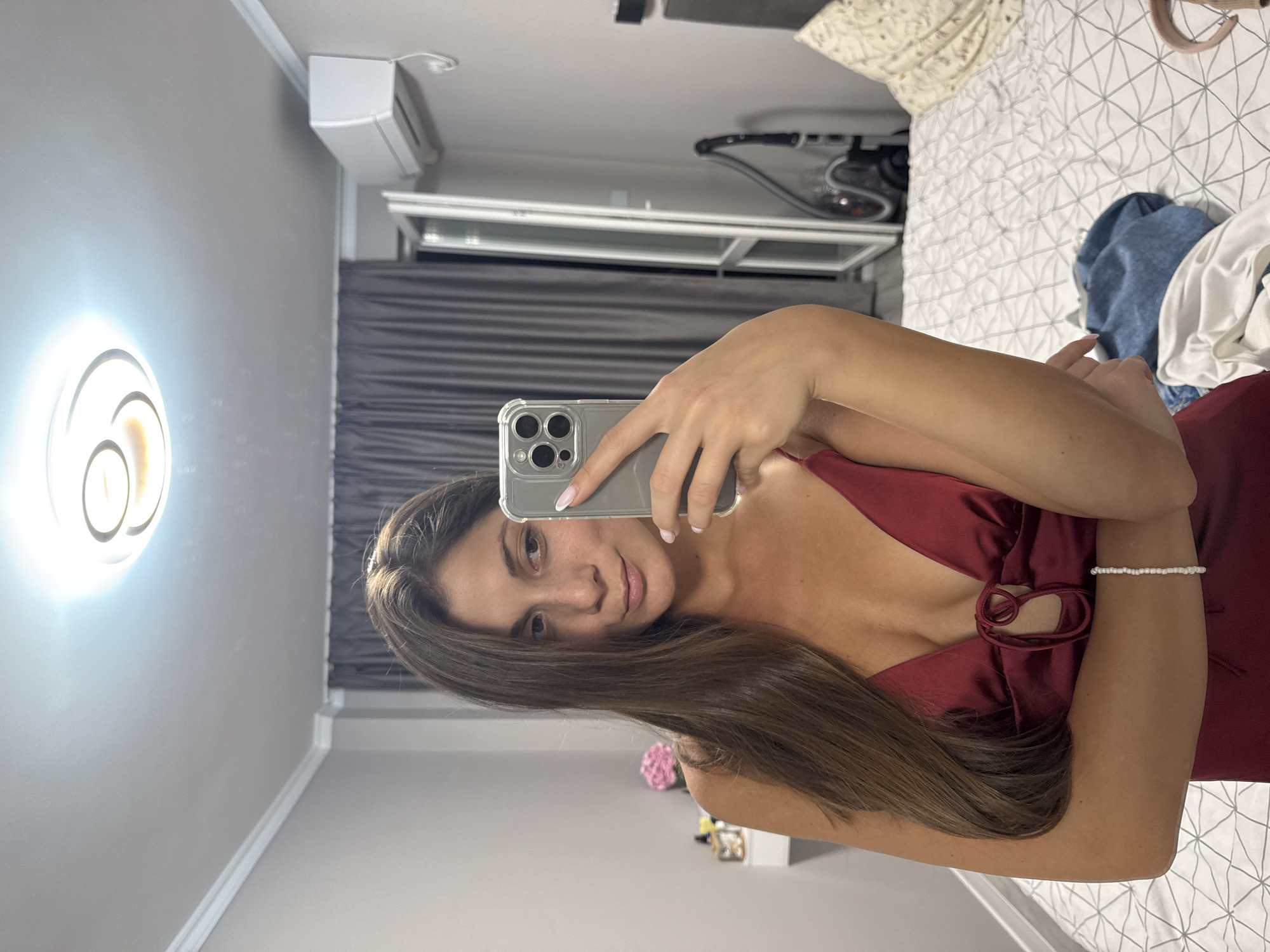 A woman is taking a selfie in a bedroom with a phone. The room is lit by an overhead circular light fixture. The woman is wearing a dark red dress. She has long, brown hair. In the background, there is a bed with a white and gray patterned comforter, gray curtains, a window, an air conditioner, and a vacuum cleaner. On a white dresser to the left, there is a bouquet of pink flowers. The scene appears to be in an apartment in Burgas, Bulgaria.