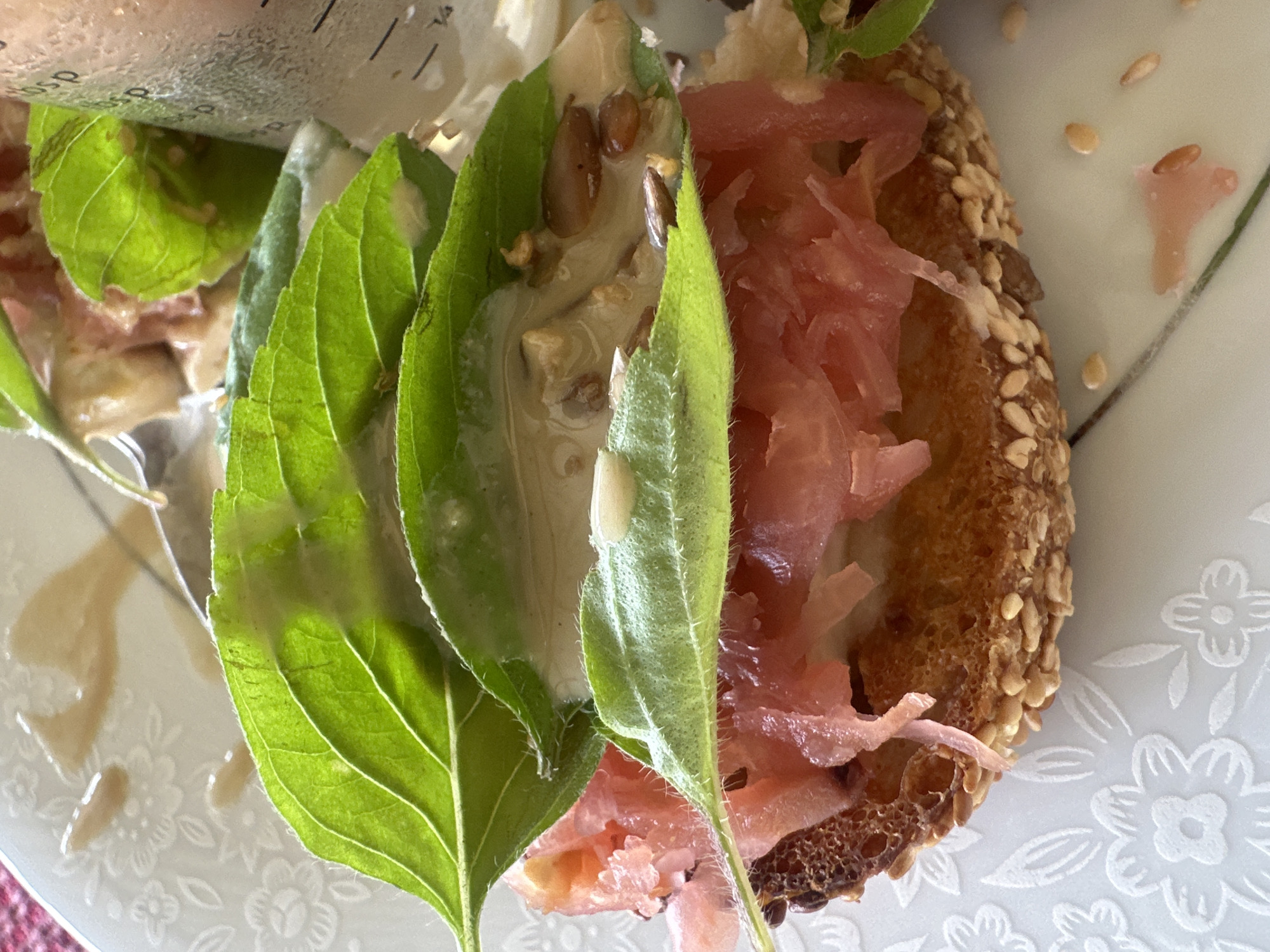The image displays a close-up of an open-faced sandwich on a white, textured plate, with a blurred background. The sandwich features a toasted, sesame-seed-crusted bread base topped with thinly sliced pink ham, green basil leaves, and a creamy sauce containing visible seeds, possibly sunflower or flax seeds. A glass with measurement markings is partially visible on the right side, suggesting it might be a beverage or part of a food preparation setup. The scene is indoors, under artificial or natural light, with the plate placed on a surface with a floral pattern. There are no people or explicit indications of the city of Thora, Australia within the image.