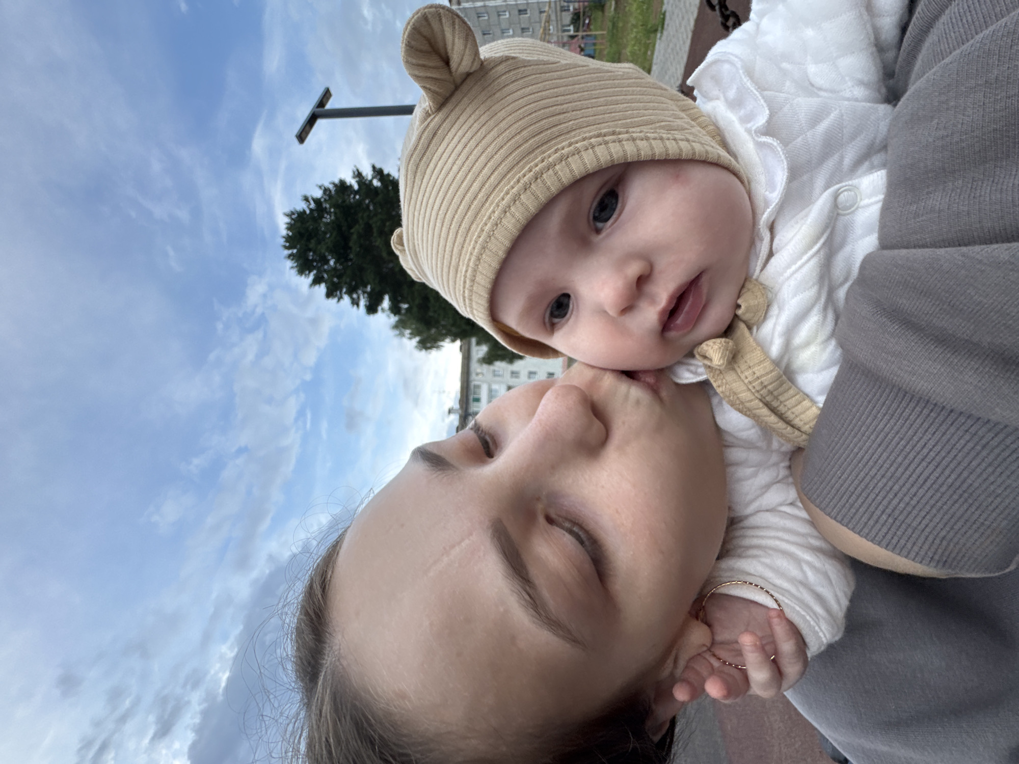 A close-up, low-angle selfie shows a young woman kissing a baby on the cheek. The woman's eyes are closed, and she is wearing a grey hooded sweatshirt and a gold hoop earring. The baby is wearing a beige ribbed knit hat with bear ears and a white quilted onesie with a ruffled collar. The background features a cloudy blue sky, a tall evergreen tree, a street lamp, and buildings in the distance, suggesting an outdoor urban setting. Based on the provided context, this scene is located in the city of Noviy, Russia.