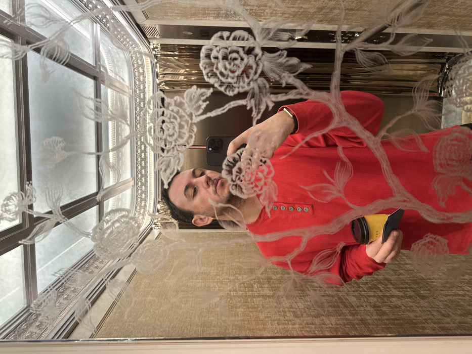 A male individual is depicted in a mirror selfie taken inside an ornate elevator, identified as likely being within The Langham London hotel at 1C Portland Place, London, United Kingdom. The elevator's interior walls feature a light-colored, textured finish, and the ceiling consists of mirrored panels with decorative metallic framing. The mirror itself is extensively etched with large, intricate floral patterns, primarily featuring peonies, branches, and foliage, which partially overlay the reflected scene.
The individual is a male with short, dark hair and light stubble. He wears a long-sleeved, ribbed red Henley shirt with four grey buttons at the collar and a single gold hoop earring in his left ear. A smartwatch with a gold-toned band is visible on his left wrist. He is holding a dark-colored smartphone, equipped with multiple camera lenses, in his right hand, positioned to capture the mirror reflection. In his left hand, he holds a yellow disposable coffee cup with a dark brown lid, supported by a dark blue/black cardboard sleeve, with a small white paper peeking from beneath the sleeve. The individual's eyes are directed towards the camera.