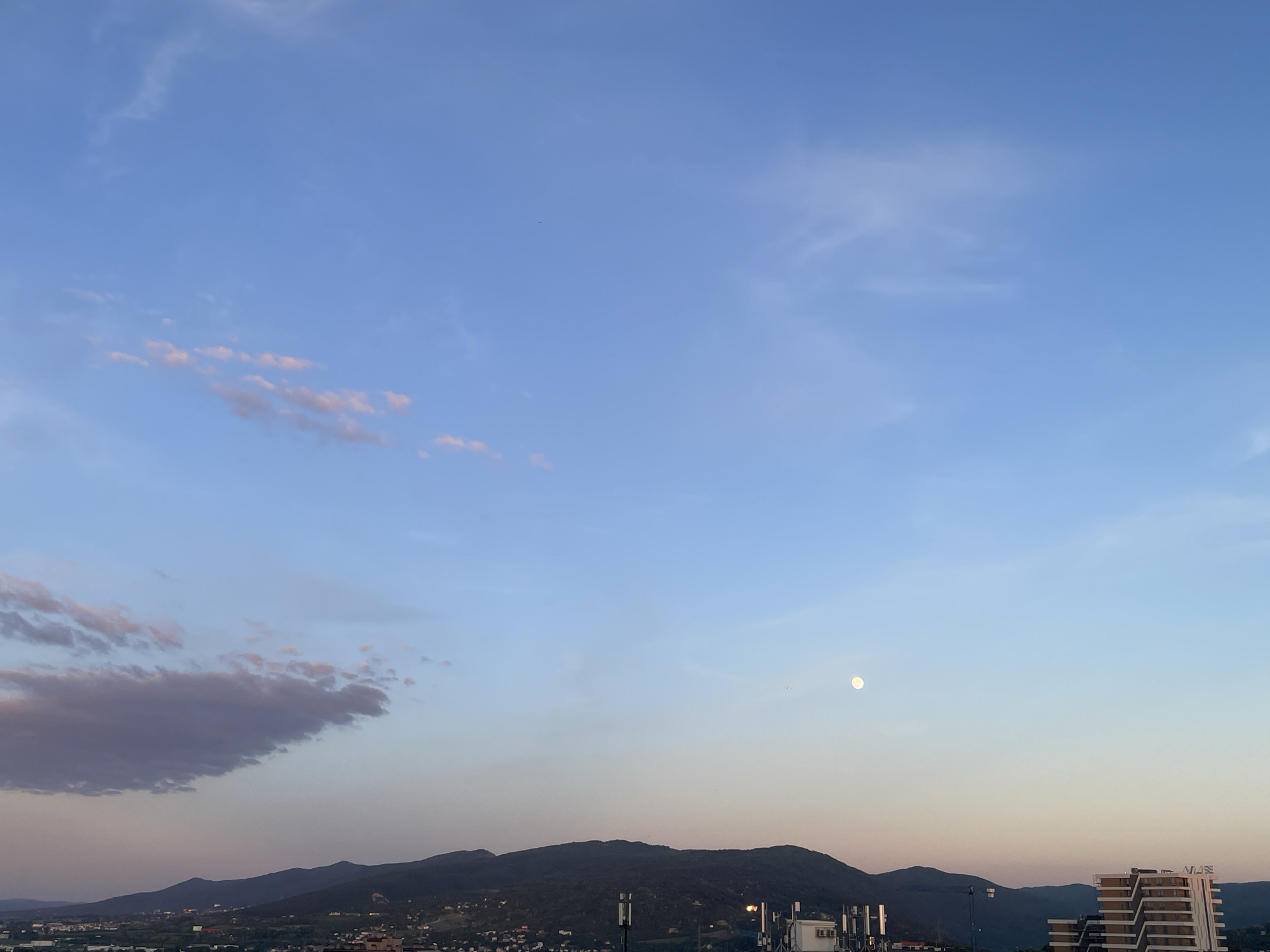 The image shows a twilight scene over Sofia, Bulgaria. In the foreground, there is a mountain range and part of the city. On the right side of the image, there is a modern building with the letters "VJS" visible on it. Also in the foreground are communications towers. In the sky, there are some clouds, and a nearly full moon. The sky is a mix of blue and orange colors.