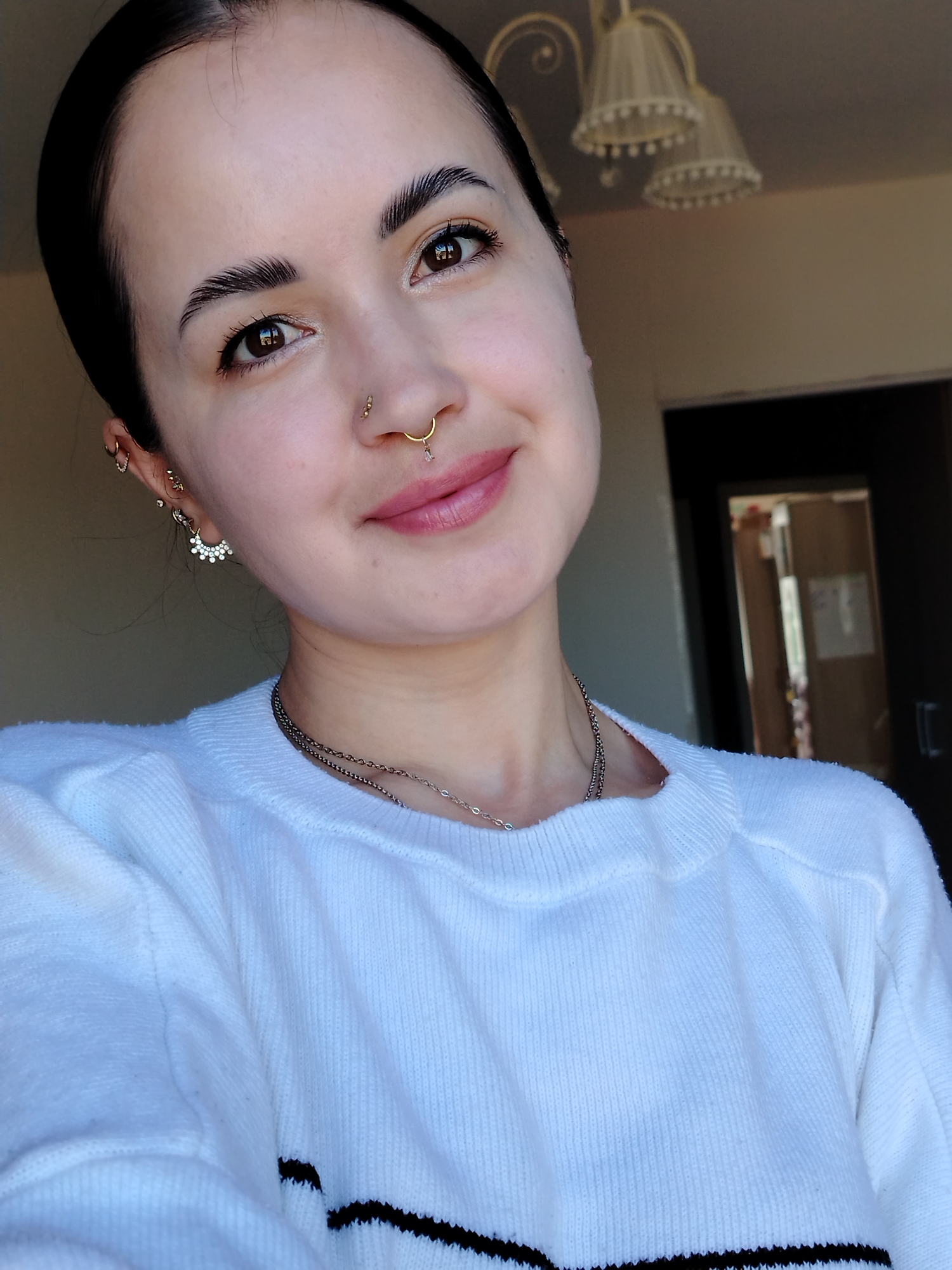 A medium-shot indoor photograph shows a woman with dark hair pulled back, brown eyes, and fair skin. She is smiling slightly, facing the camera at a tilted angle. She has multiple piercings in her left ear and nose. She wears a white knit sweater with a black stripe near the bottom. Two necklaces are visible around her neck. The background shows a door and a light fixture with multiple lampshades. The location is likely indoors and cannot be verified to be in Noviy, Russia.
