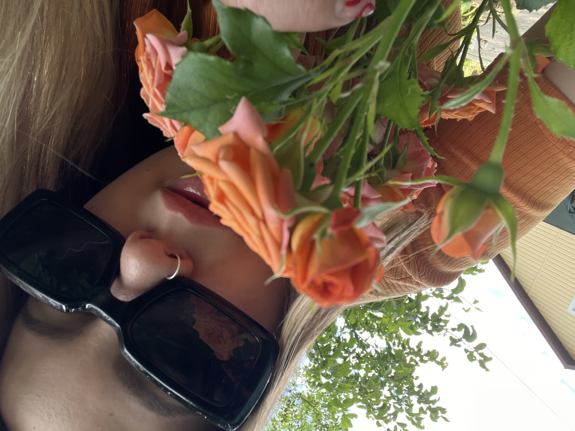 This is a close-up, portrait-oriented photograph taken outdoors. The primary subject is a person's face, largely obscured by large, black, rectangular sunglasses. Only a portion of the forehead, nose with a silver hoop piercing, lips, and chin are visible. The person has fair skin and blonde hair that frames the visible parts of their face. They are holding a bouquet of small, pink and orange roses. The hand holding the flowers is visible at the top of the frame, with manicured nails painted with red and white accents. The person is wearing an orange, ribbed garment, possibly a sweater or top.
The background suggests an outdoor setting, with green foliage from trees and a pale sky with some hints of clouds. A portion of a building with a yellow, corrugated roof and brown trim is also visible on the right side of the frame. The lighting indicates it is daytime, likely bright, possibly sunny given the high contrast and reflections in the sunglasses. There is no visible text in the image. The overall impression is casual and summery, with a focus on the flowers and a sense of relaxed enjoyment of the outdoors.