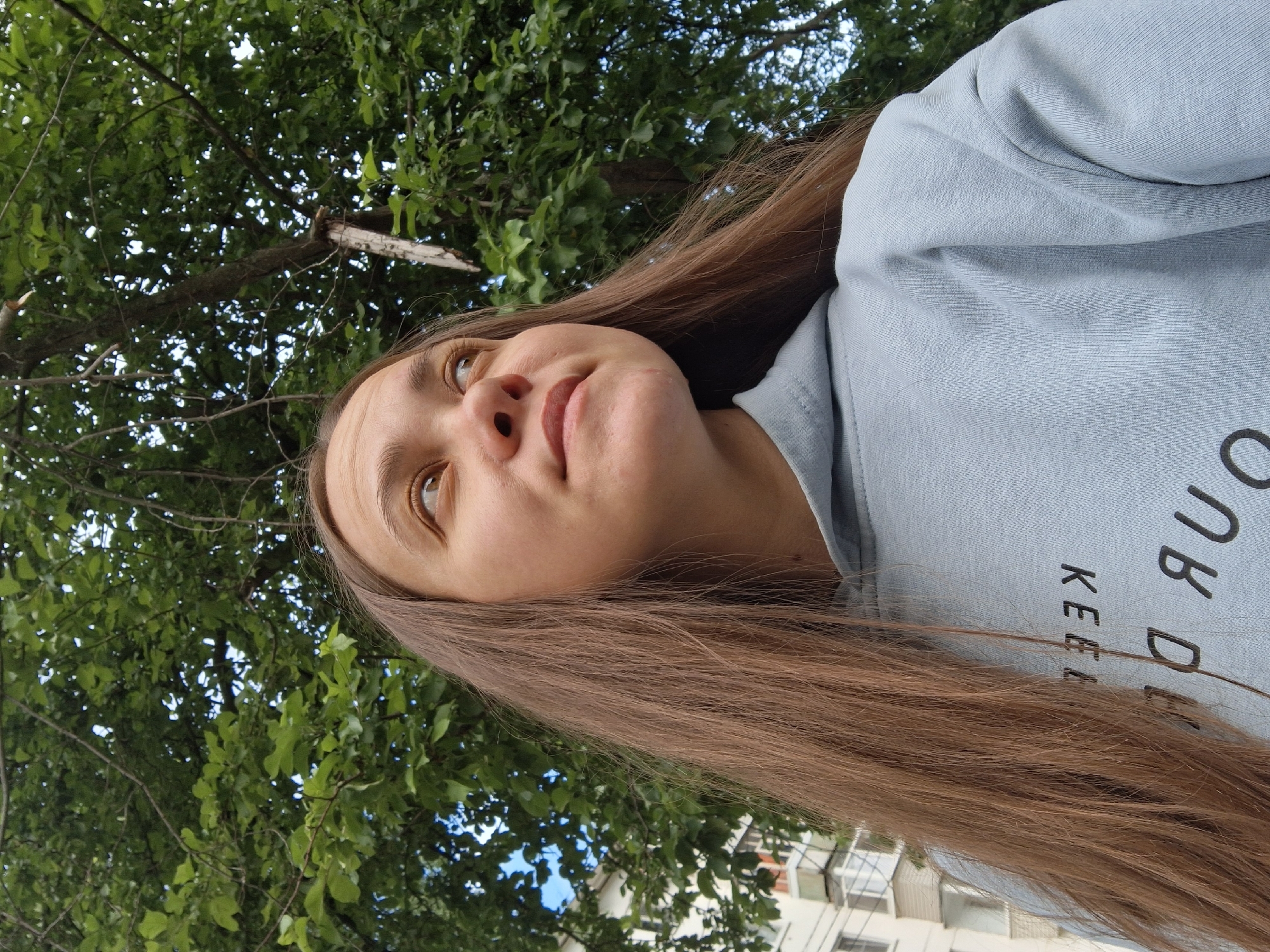 A low-angle, close-up portrait features a person, likely a young woman, with long, straight, light brown hair flowing down her back and shoulder. Her head is tilted slightly back, and she is looking upwards with a contemplative expression and a subtle smile. Her eyes are a striking light brown or amber color.
She is wearing a light blue or light grey hooded sweatshirt. Visible text on the front of her sweatshirt includes the words "KEEP" and "DON'T", possibly part of a larger phrase.
The background is dominated by a dense canopy of vibrant green tree leaves, suggesting an outdoor setting under a tree. Patches of bright blue sky are visible through the foliage, indicating clear weather and daytime. In the far distance, a blurry, light-colored multi-story building with visible windows can be discerned, hinting at an urban or residential environment.
The scene conveys a calm and observant mood, as if the person is pausing to appreciate her surroundings. The overall impression is a casual moment captured in a green space within Chaikovsky, Russia.