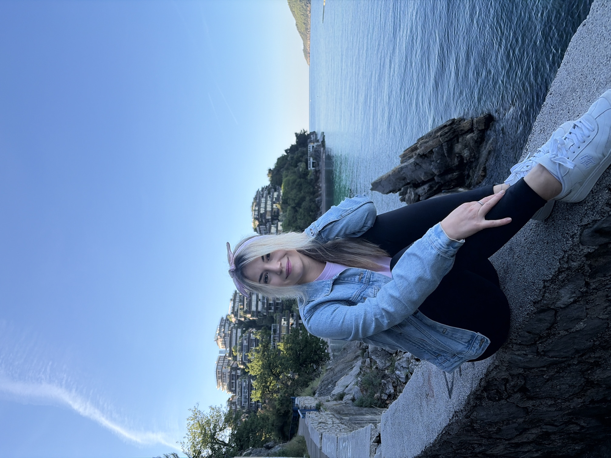 A young woman with blonde hair and a purple headband sits on a stone wall with her legs crossed. She wears a denim jacket, a light purple shirt, and black leggings. She has white sneakers on her feet. Behind her is a view of the Adriatic Sea, with rocky outcrops and buildings visible along the coastline. The image is taken in Budva, Montenegro, along the coastal path near the Avala Resort. The sky is a clear, light blue.