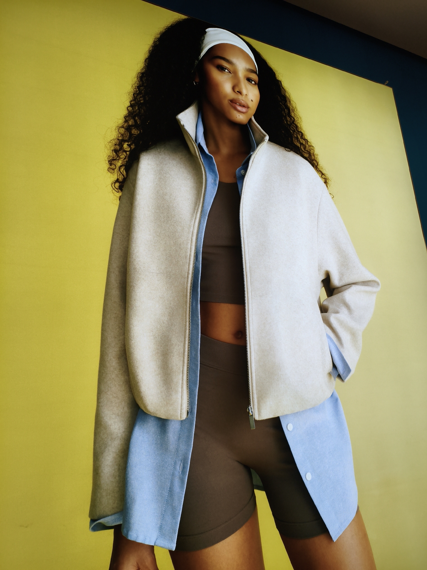 A woman with dark, curly hair and a white headband stands against a yellow backdrop. She is wearing a beige zippered jacket over a brown crop top and brown shorts. A light blue collared shirt is layered beneath the jacket, with its collar and sleeves visible. The background features a split in color, with a dark blue section above the yellow. The image appears to be a studio portrait. There is no indication of any specific location in Sandwell, United Kingdom.