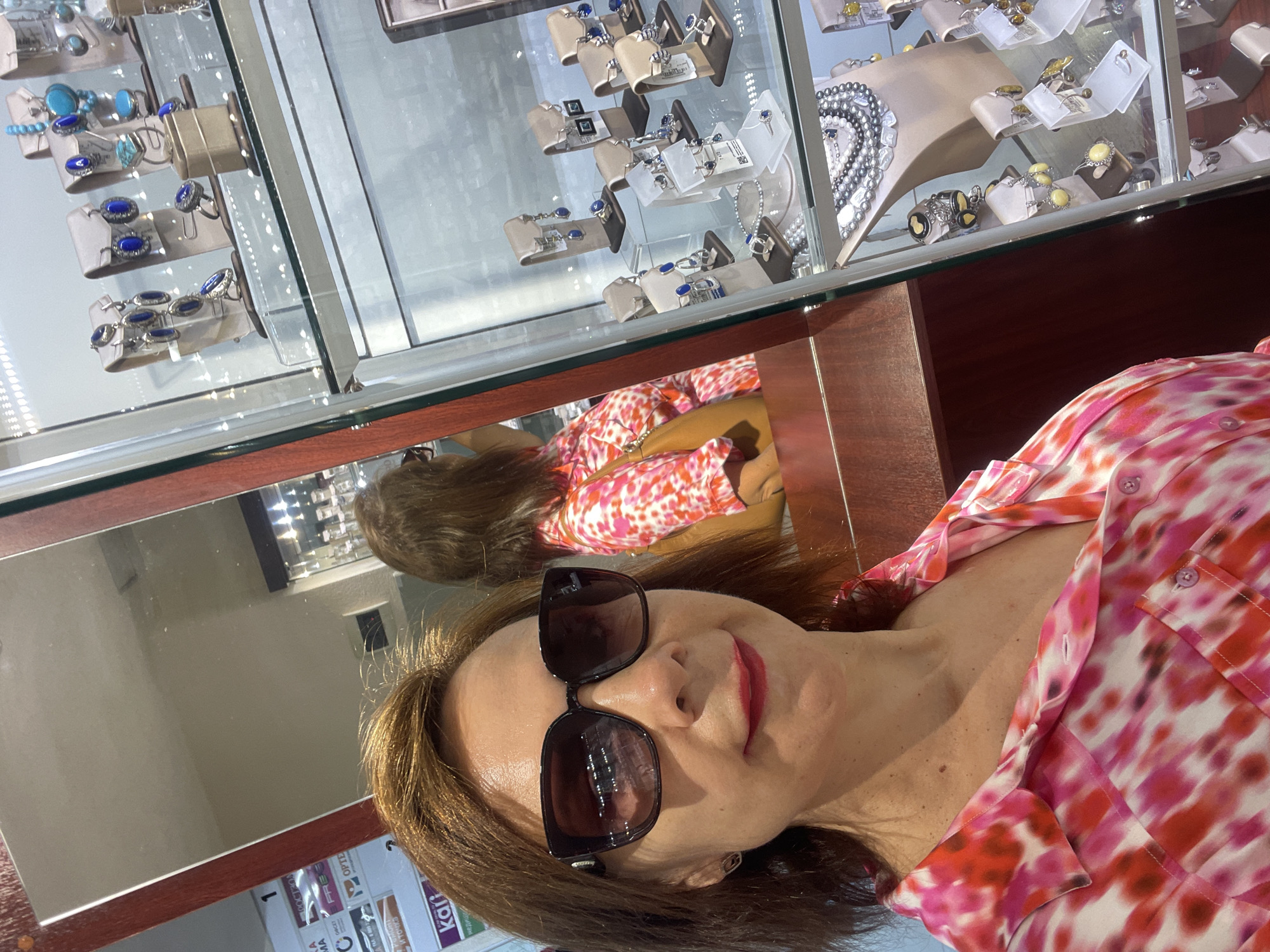 A woman with reddish-brown hair and sunglasses is taking a selfie in a jewelry store. She is wearing a pink and white tie-dye shirt and has red lipstick on. A jewelry display case is visible to her right, filled with necklaces, earrings, and rings. A reflection of another woman in a pink and white patterned top is visible in a mirror behind the selfie-taker. The store appears to be located in Sochi, Russia, as indicated by the Cyrillic text on posters in the background.