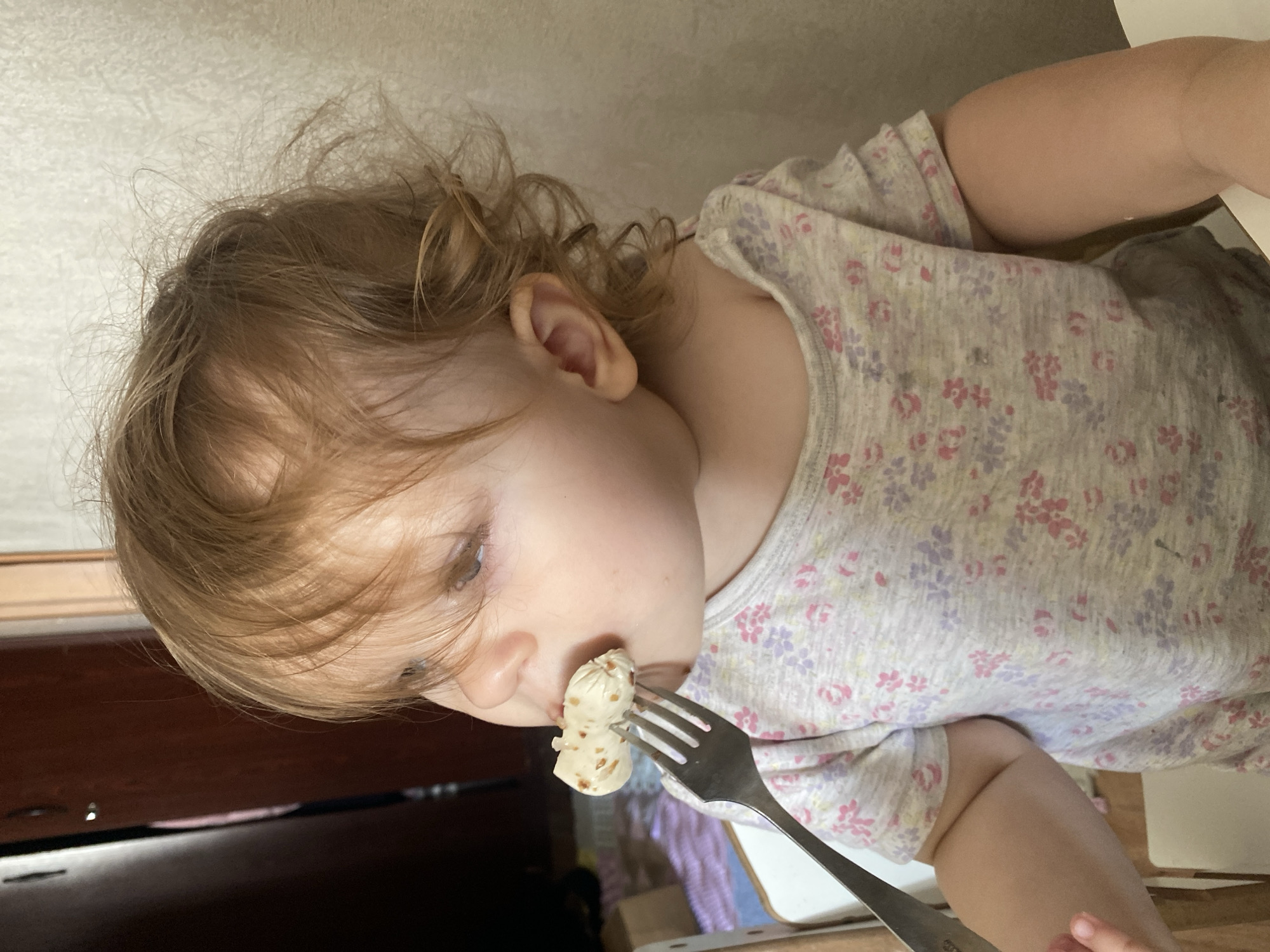 A young child is eating a piece of food skewered on a fork. The child has light skin, brown hair, and is wearing a floral-patterned shirt. The child is looking down at the food on the fork, which is being held up to their mouth. The background is blurry, but shows a table and other furniture. The location in the city of Unknown, Unknown cannot be determined.