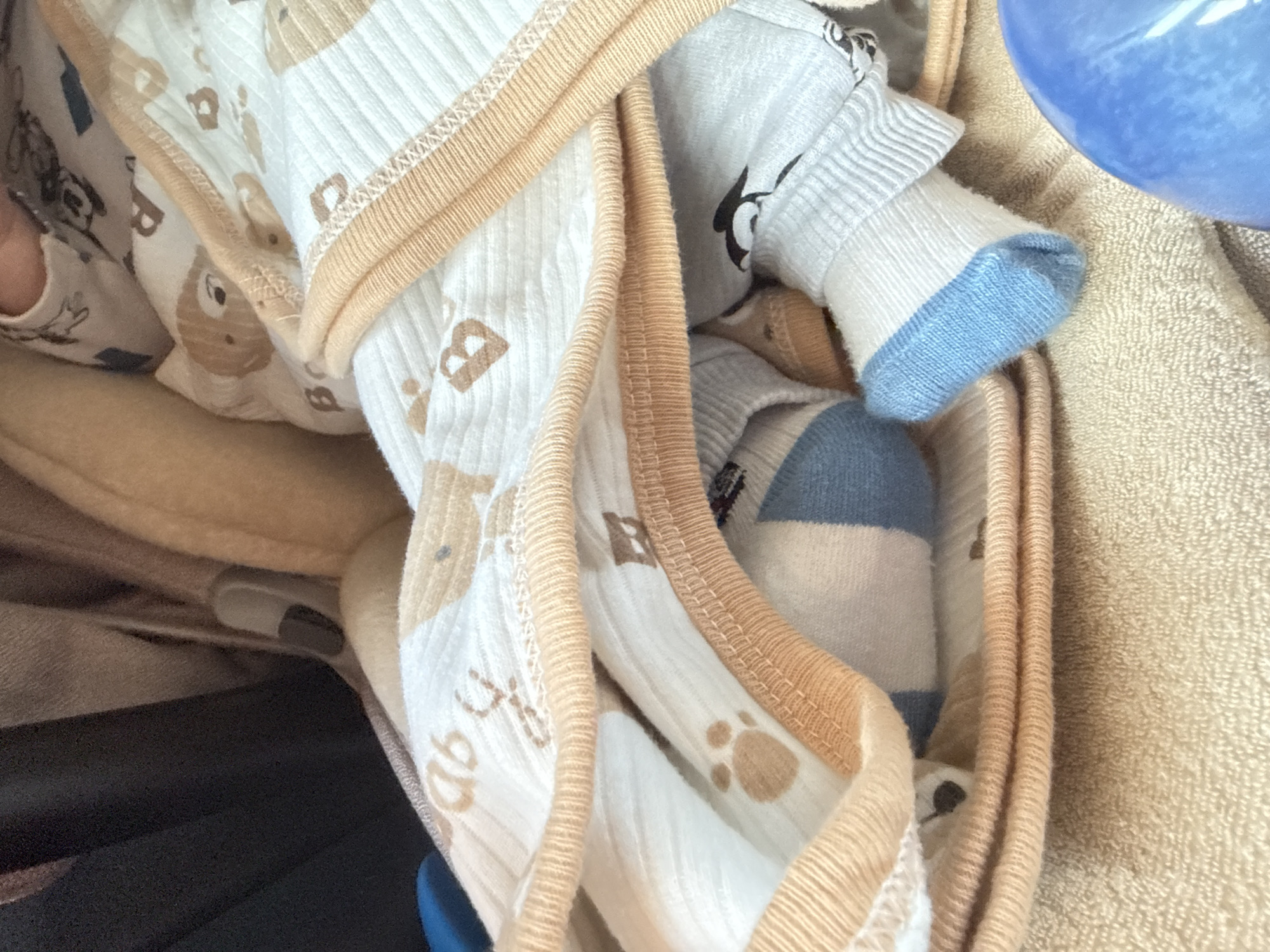 A close-up, high-angle shot captures a collection of baby clothes and textiles. The primary focus is on several pieces of light-colored baby apparel, folded or stacked together. Many of the garments are made from a white, ribbed fabric featuring a pattern of light brown cartoon bears, paw prints, and what appears to be the letter "b" or "ab" repeated. These clothes have beige or light brown trim.
Interspersed with these items are pieces of light blue and white baby clothing, including a light blue sock with a white body, the blue toe clearly visible. To the right, a textured, light beige or tan towel or blanket is visible. At the top right, a smooth, bright blue, curved object, possibly part of a plastic cup or toy, is partially visible. The bottom left shows a darker, possibly black or dark blue fabric, which could be part of a bag, container, or furniture. In the far left, a small portion of a human finger or hand is visible, suggesting someone is interacting with or arranging the items. The overall scene conveys a sense of domestic activity, likely involving the sorting or preparation of baby items in an indoor setting in Rogosh, Bulgaria.