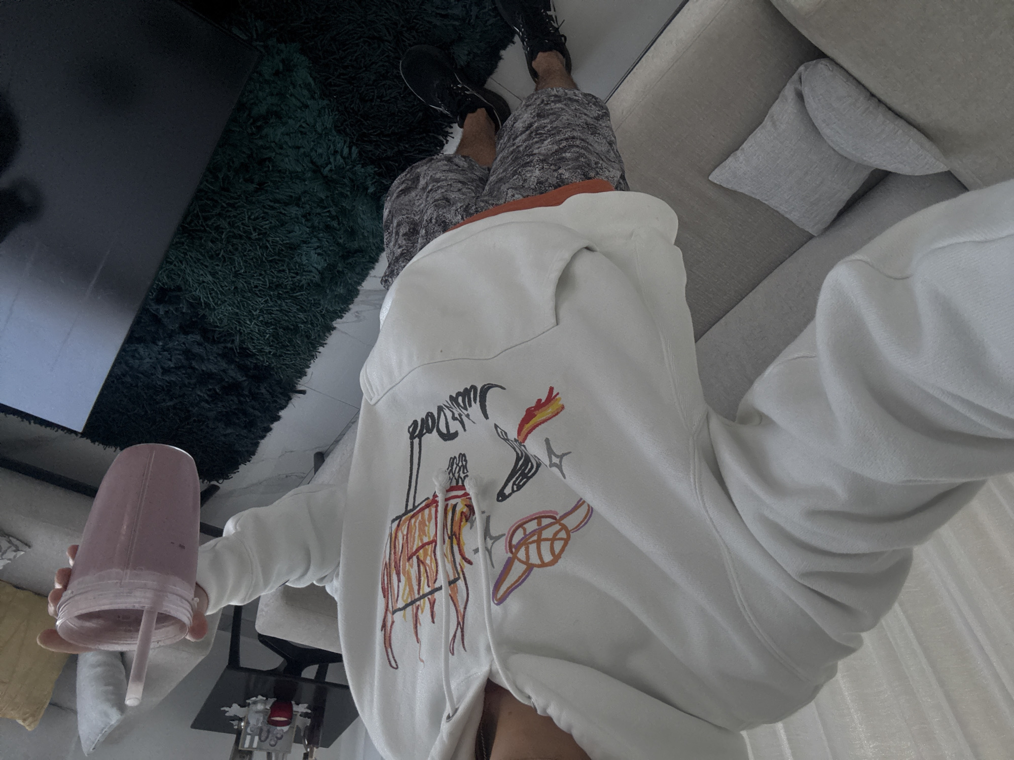 The image, taken from a high-angle perspective and rotated, depicts a person from the chest down, holding a translucent pink-purple smoothie cup with a straw. The individual is wearing a white hooded sweatshirt adorned with colorful, hand-drawn style graphics. Visible text on the hoodie reads "Just Don" in a stylized script, accompanied by illustrations of a barbecue grill with flames, a flaming unicorn-like creature, and a basketball with rings. Below the hoodie, the person wears camouflage-patterned shorts and black sneakers.
The setting appears to be an indoor living space, consistent with a residence in San Bernardino Tlaxcalancingo, Mexico. A dark green shaggy rug lies on a light-colored floor, partially beneath a dark, rectangular piece of furniture. To the person's right, a light-colored fabric sofa with a grey accent pillow is visible. White curtains or blinds can be seen in the background. The lighting suggests it is daytime. The activity involves holding a beverage, possibly during a moment of relaxation. No specific emotions are conveyed as the person's face is not shown.