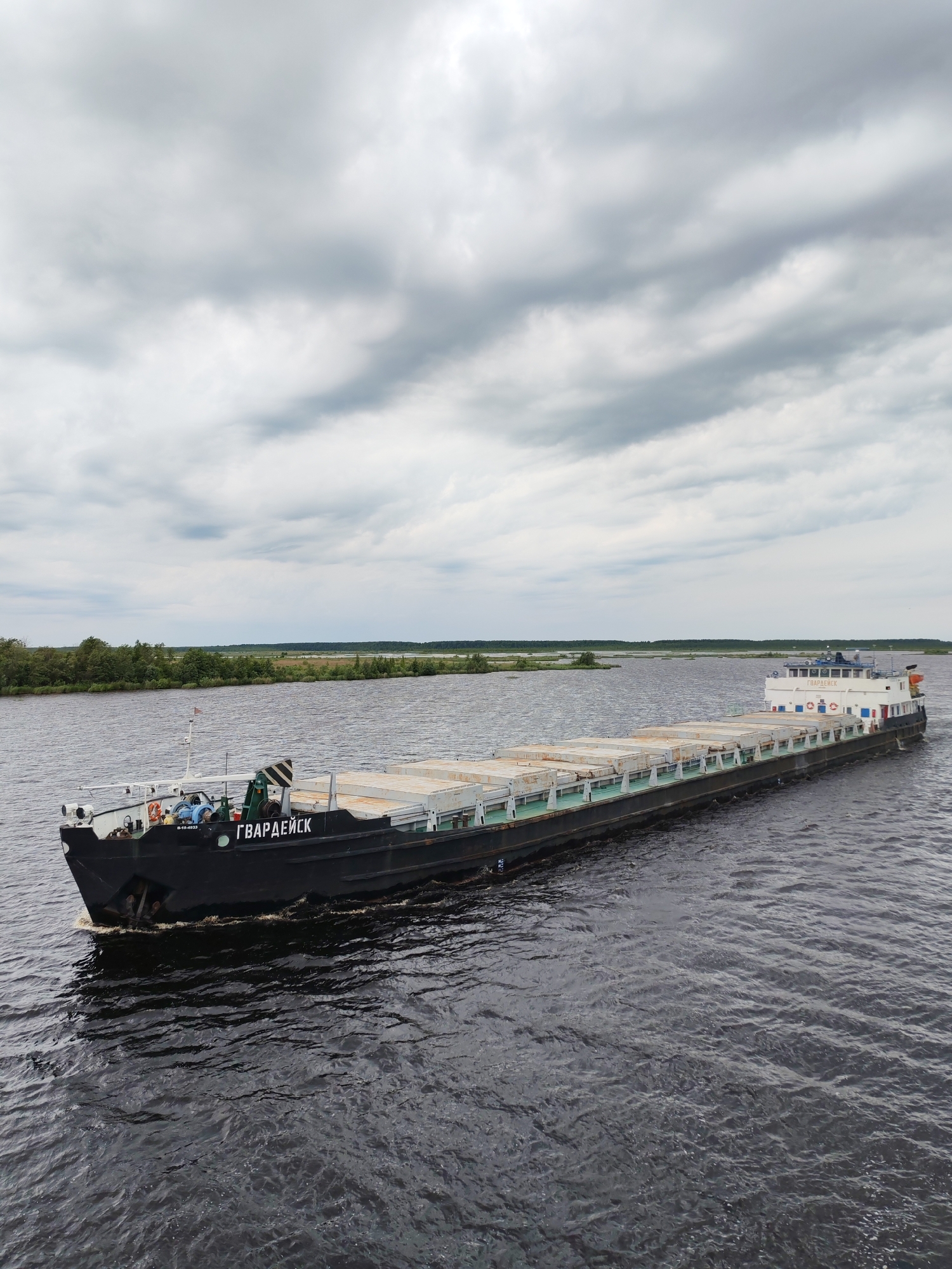 A barge named "ГВАРДЕЙСК" sails on a dark, choppy river under a cloudy sky. The barge, which appears to be a cargo barge, is laden with rectangular, pale-colored containers. The barge is black with a white superstructure at the stern, which features windows and life preservers. A crane is visible on the deck. The riverbanks in the background are lined with green trees. The location is identified as Novokemskii, Russia.