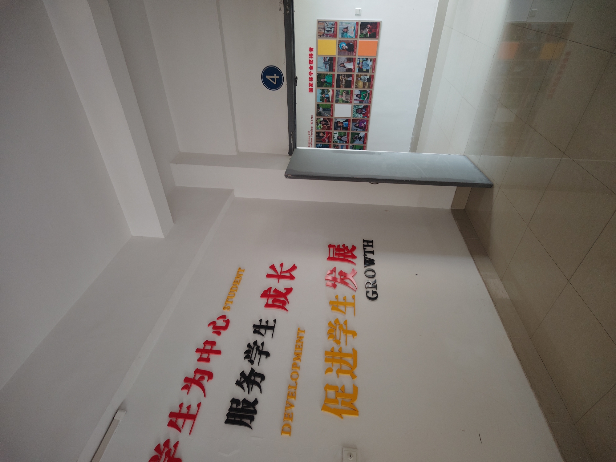 The image shows an indoor hallway in what appears to be an educational institution in Chongqing High-tech Zone, China. The walls are white. On one wall, there is a large sign with Chinese characters and English translations. The text reads: "学生为中心 STUDENT 服务学生成长 DEVELOPMENT 促进学生发展 GROWTH." To the right of the hallway, a sign with a grid of pictures is visible, with the number 4 on a blue circle to the left of it. The floor has light brown tiles. The ceiling appears to have layered white beams.
