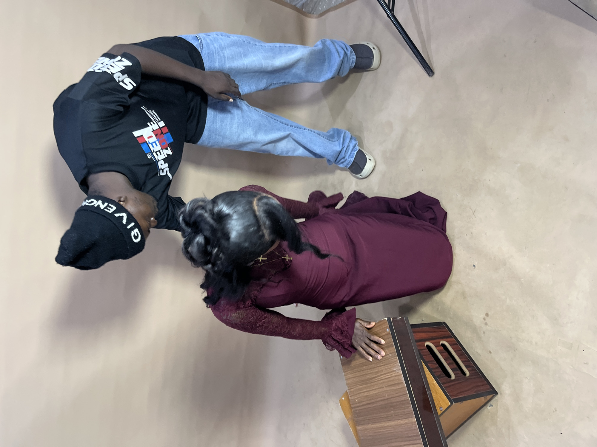 In an indoor studio setting with a plain, light brown backdrop, a Black male and a Black female are the subjects. The male, wearing a black beanie with white text and a black t-shirt with a graphic design that includes the words "SPEED ZONE," is leaning over and looking down at the female. He is also wearing light blue jeans and grey shoes. The female is kneeling on the floor, facing away from the camera and slightly to the left. She is wearing a long, maroon dress with lace sleeves that have ruffled cuffs and a gold cross necklace. Her hair is styled in an elaborate updo. To her left is a wooden object, possibly a small table or stand, with visible drawers. The floor is a textured, light beige material with some scattered dark marks.