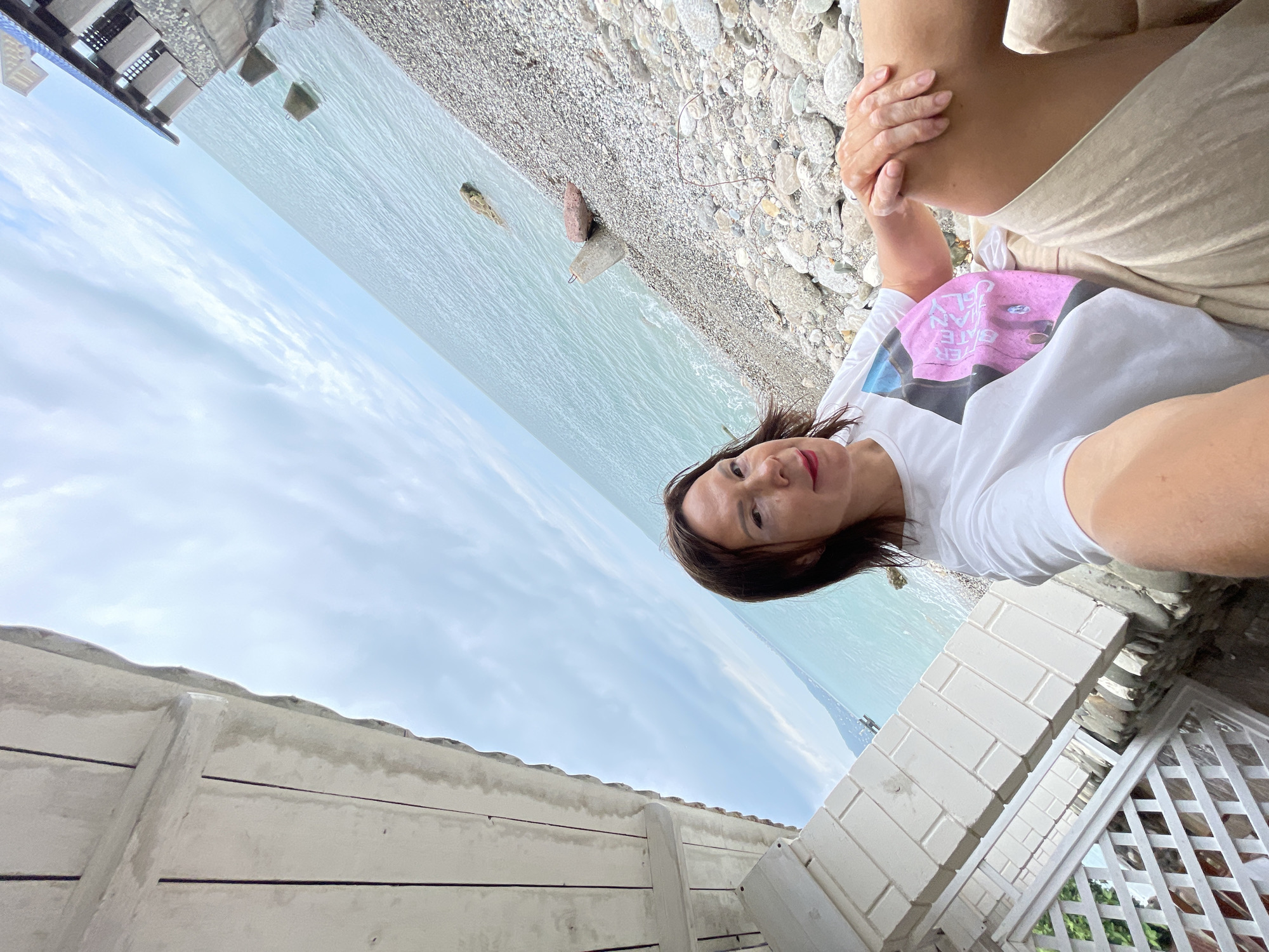 A woman is taking a selfie on a pebble beach in Abaata, Abkhazia. She is wearing a white t-shirt with a graphic print and tan shorts. She is sitting down, with her left hand resting on her left knee. The background shows the sea and a cloudy sky. There are some concrete blocks in the water and on the beach. Part of a white structure with a lattice design is visible on the left side of the image.