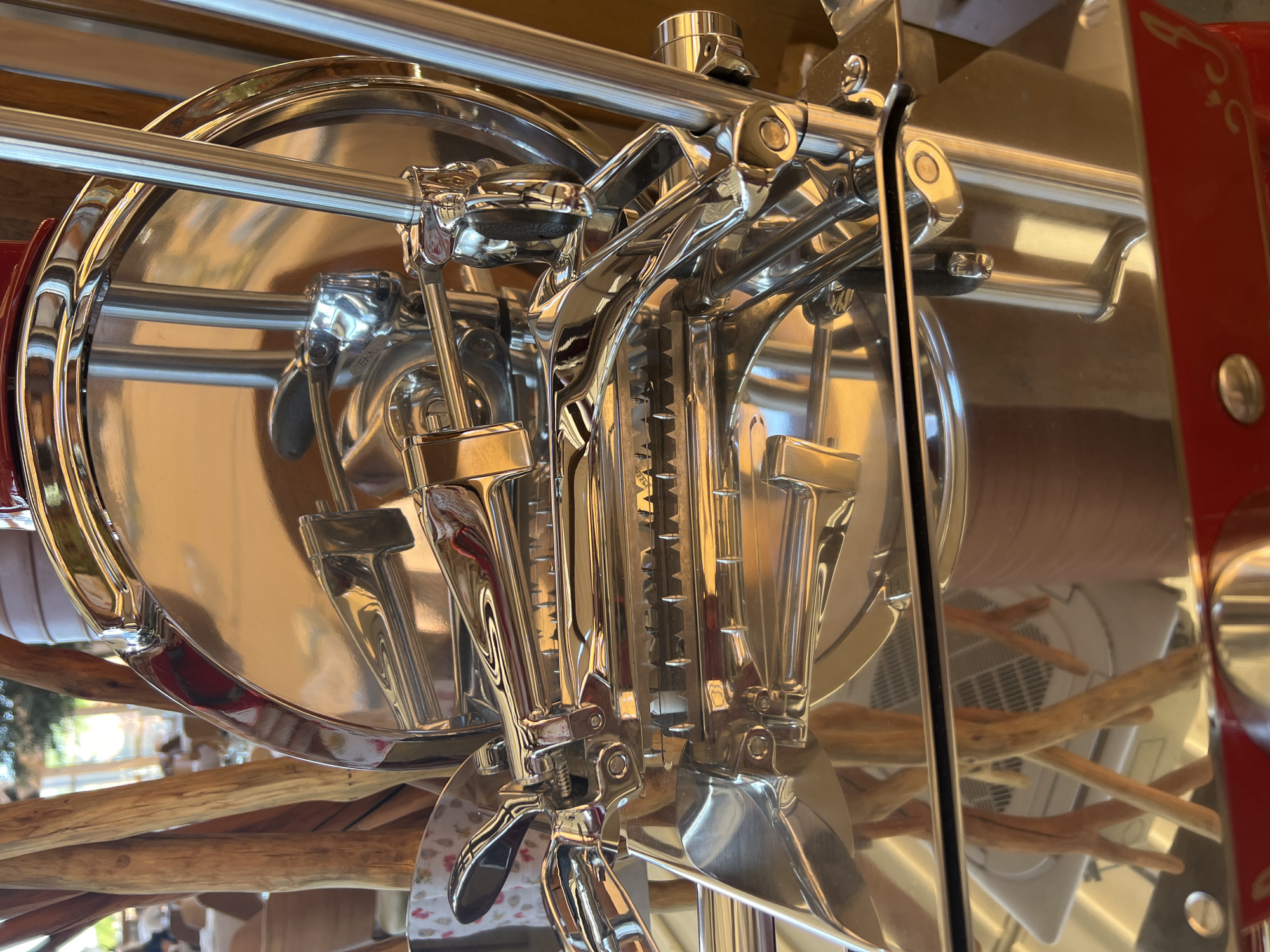 The image shows a close-up of a polished chrome-plated meat slicer, likely in a commercial setting. The slicer has a large circular blade, a carriage mechanism, and several levers and handles, all reflecting the surrounding environment. The background is blurred, but appears to be an indoor space with natural wood elements and possibly tables and chairs, suggesting a restaurant or deli. There are no people visible in the immediate foreground. The image's context suggests it was taken in Kovačko Polje, Montenegro, though the specific location within the city is not discernable from the image itself.