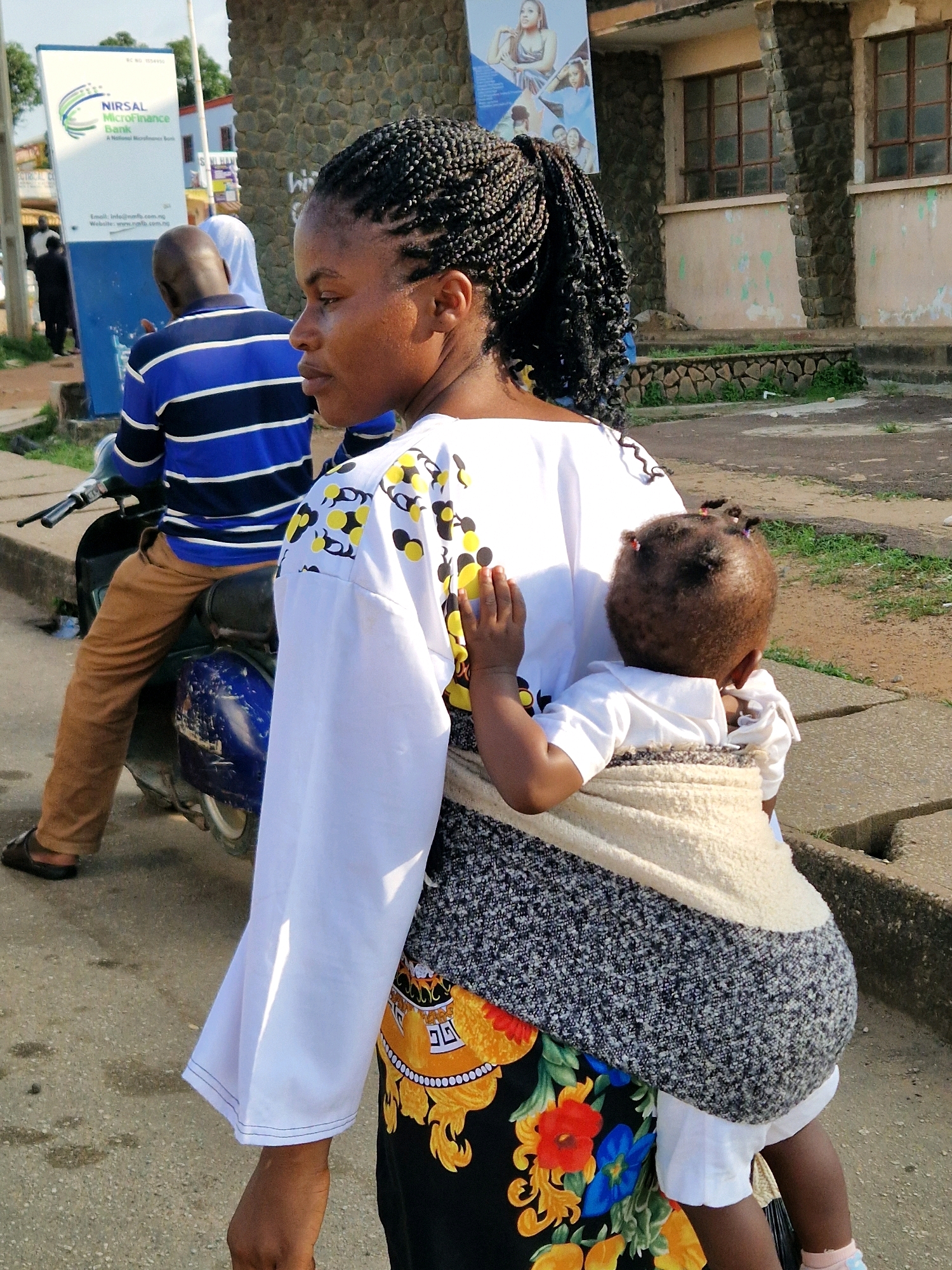 A young Black woman with braided hair walks away from the camera, carrying a baby on her back in a fabric sling. The woman is wearing a white shirt with a yellow and black circular pattern on the sleeves and a black skirt with a colorful floral design. The baby, facing away from the camera, is wrapped in a cream-colored fabric sling. In the background, a man wearing a blue and white striped shirt and brown pants sits on a blue motorcycle. To the left, a white sign with green text reads "NIRSAL MicroFinance Bank." The scene appears to be outdoors on a paved street with a stone wall and building in the background.