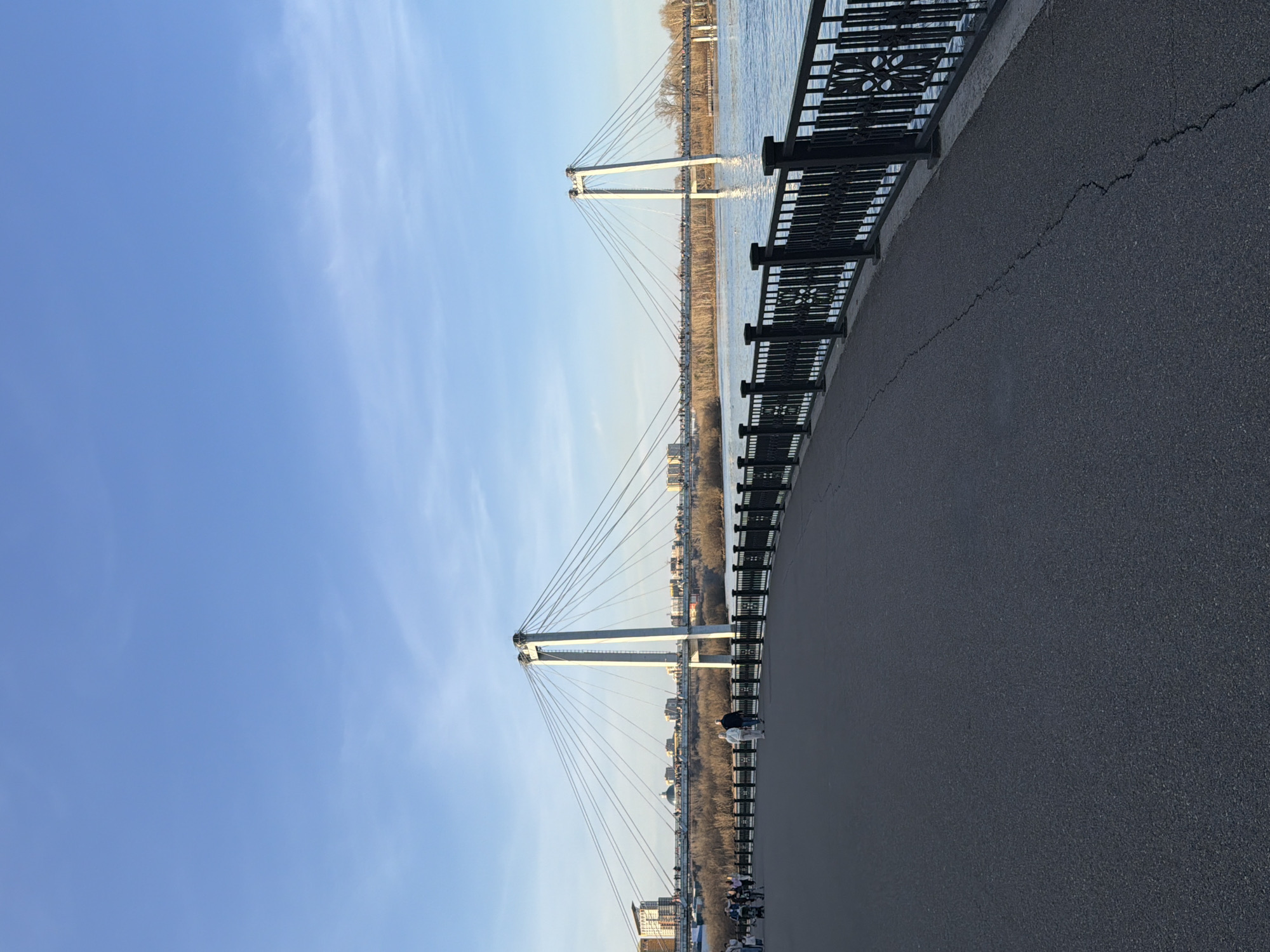 The image shows a pedestrian walkway alongside the Yenisey River in Krasnoyarsk, Russia, featuring the Vinogradovsky Bridge. Two white pylons with suspension cables are visible, extending over the water. A black metal railing separates the walkway from the river. People are seen walking and relaxing on the pavement. The sky is clear and blue. The asphalt walkway has a visible crack running along its edge. Buildings and trees line the opposite bank of the river.