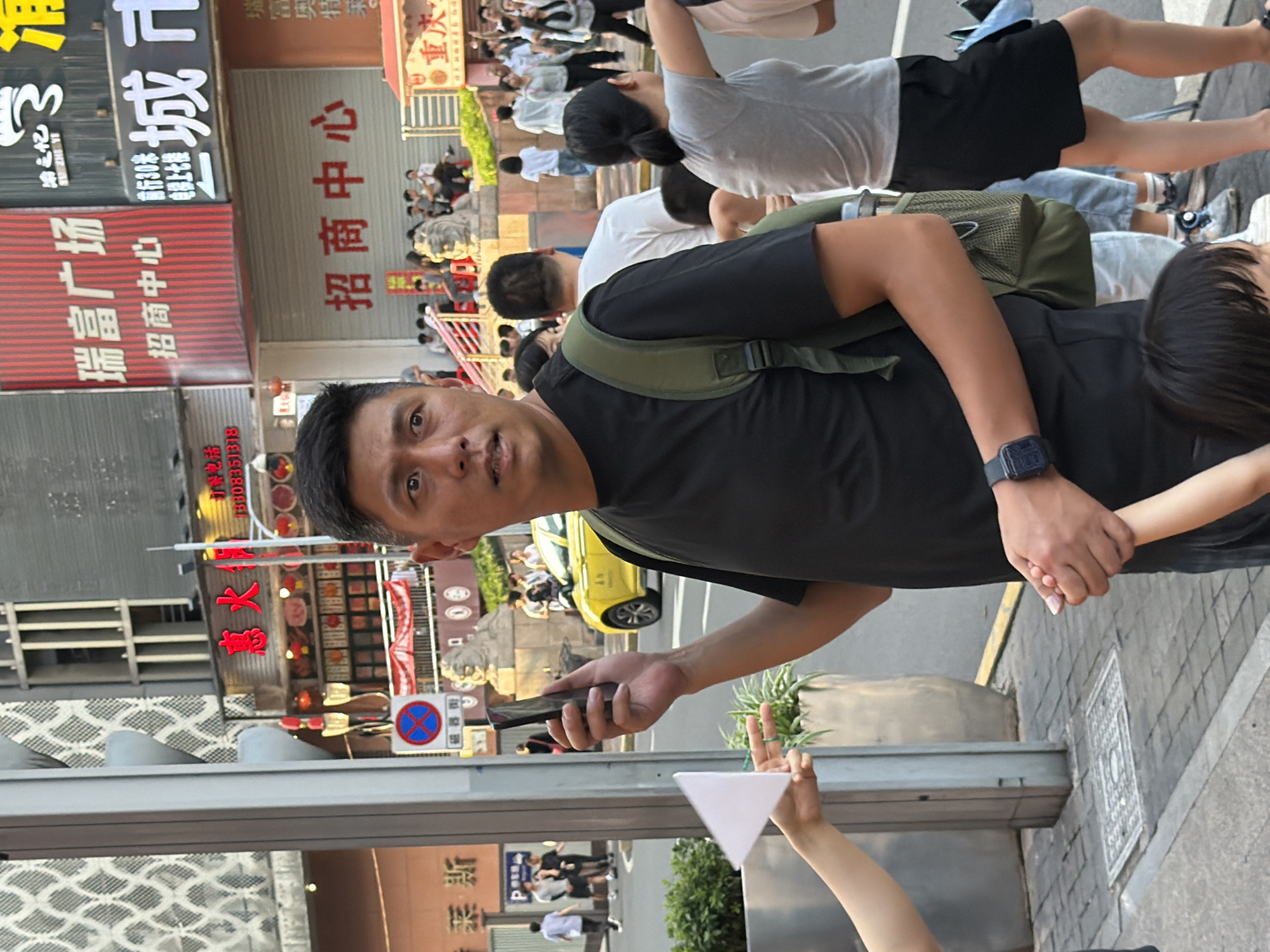 A man in a black t-shirt and a green backpack holds a smartphone in his right hand, looking towards his right. His left hand is extended, holding the hand of a child whose head is visible at the bottom of the frame. A woman in a grey t-shirt and black skirt stands to the man's right, facing away from the camera. Other pedestrians are visible in the background, as well as a yellow car. The scene appears to be a city street in Yuzhong District, China, with buildings and signs in Chinese characters. There is a traffic sign with a red circle and a blue background with a red diagonal line and a red horizontal line.