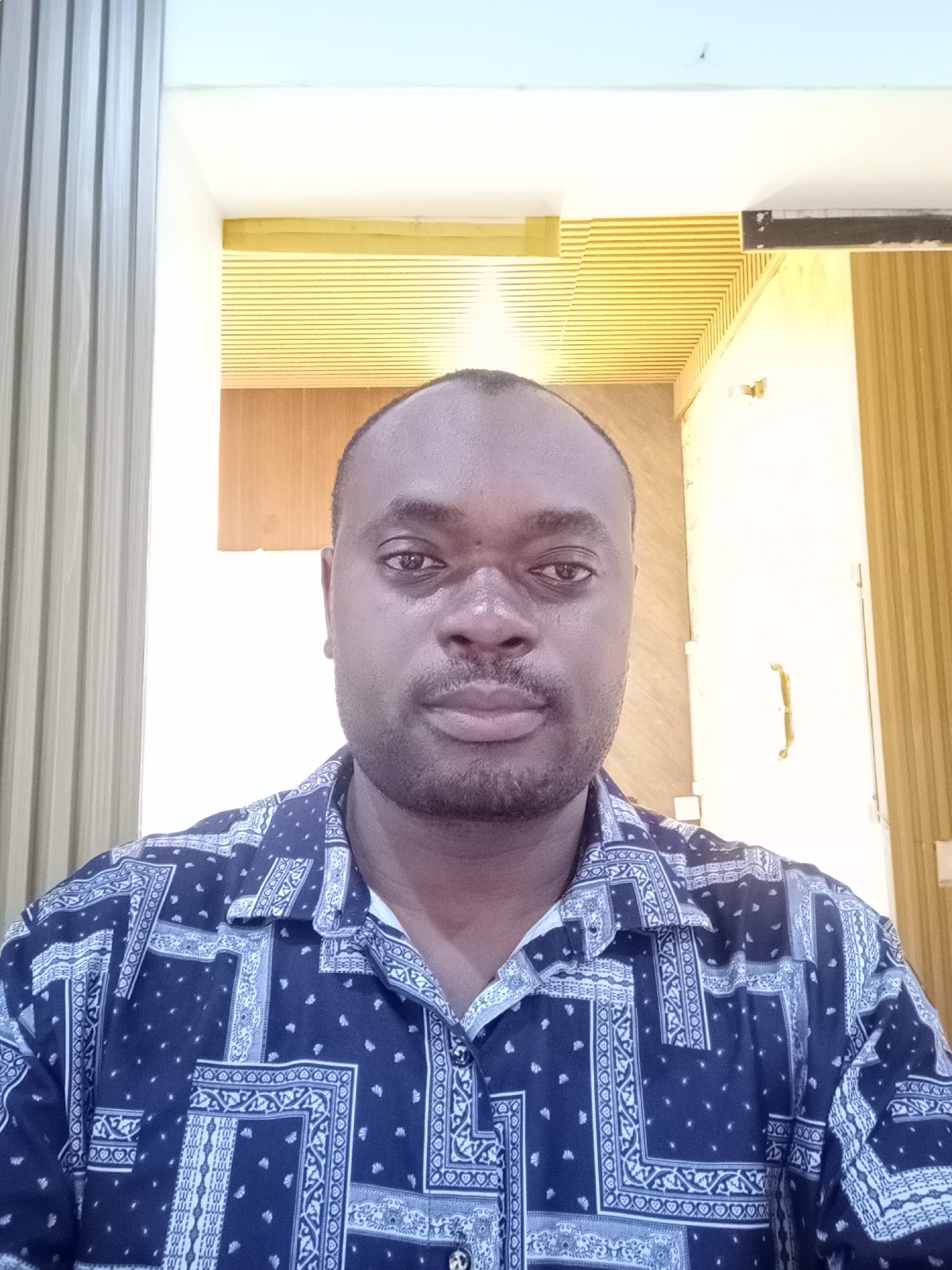 A medium-shot photograph depicts a dark-skinned man centered in the frame, wearing a dark blue patterned shirt with a button-down collar. The man has a shaved head and a short beard. The background consists of a doorframe or opening. The interior space has alternating light and dark vertical paneling to the left, while a glimpse into a room with a golden ceiling and light wooden door is visible through the opening. The location is consistent with an indoor space in Gwarinpa, Nigeria.