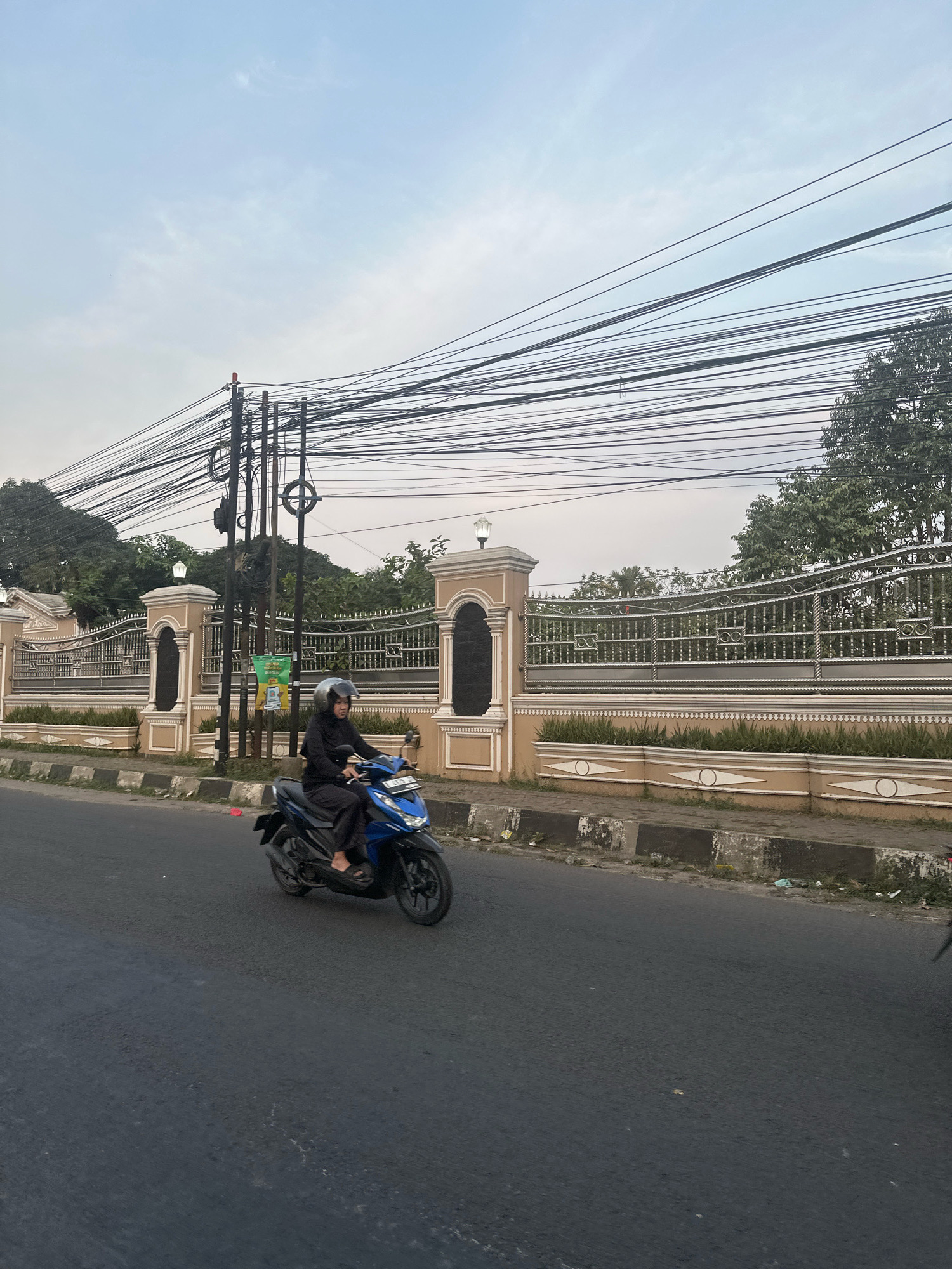 In the city of Saruni, Indonesia, a person wearing a black hijab and black clothing rides a blue scooter on an asphalt road. The scooter has a license plate with characters that are illegible due to the image quality. The rider is wearing a grey helmet. Behind the rider, a beige-colored fence with ornate metalwork borders a property lined with trees. A utility pole with a dense network of black wires is situated near the fence. The sky is overcast with a pale blue hue. The road is bordered by a concrete curb and a strip of land with sparse vegetation and debris. The overall scene depicts a typical urban streetscape in Indonesia.