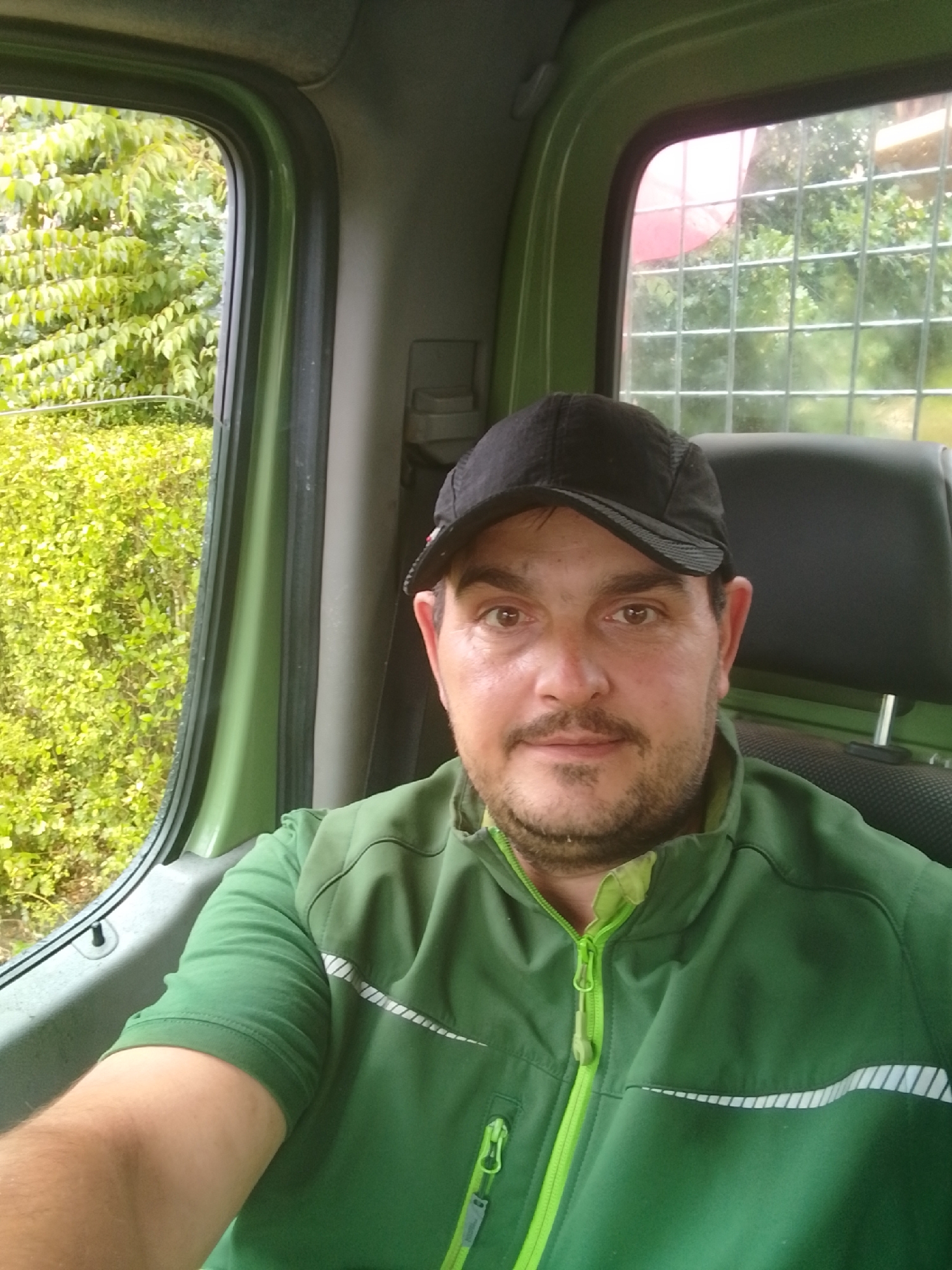 A man wearing a black baseball cap and a green zip-up jacket is inside a vehicle. He is looking at the camera and appears to be taking a selfie. The interior of the vehicle is green, and there is a black headrest visible behind the man. Outside the vehicle, through the windows, lush green foliage can be seen. The lighting suggests it is daytime. The image does not provide any specific details to determine its location within Hamburg, Germany.