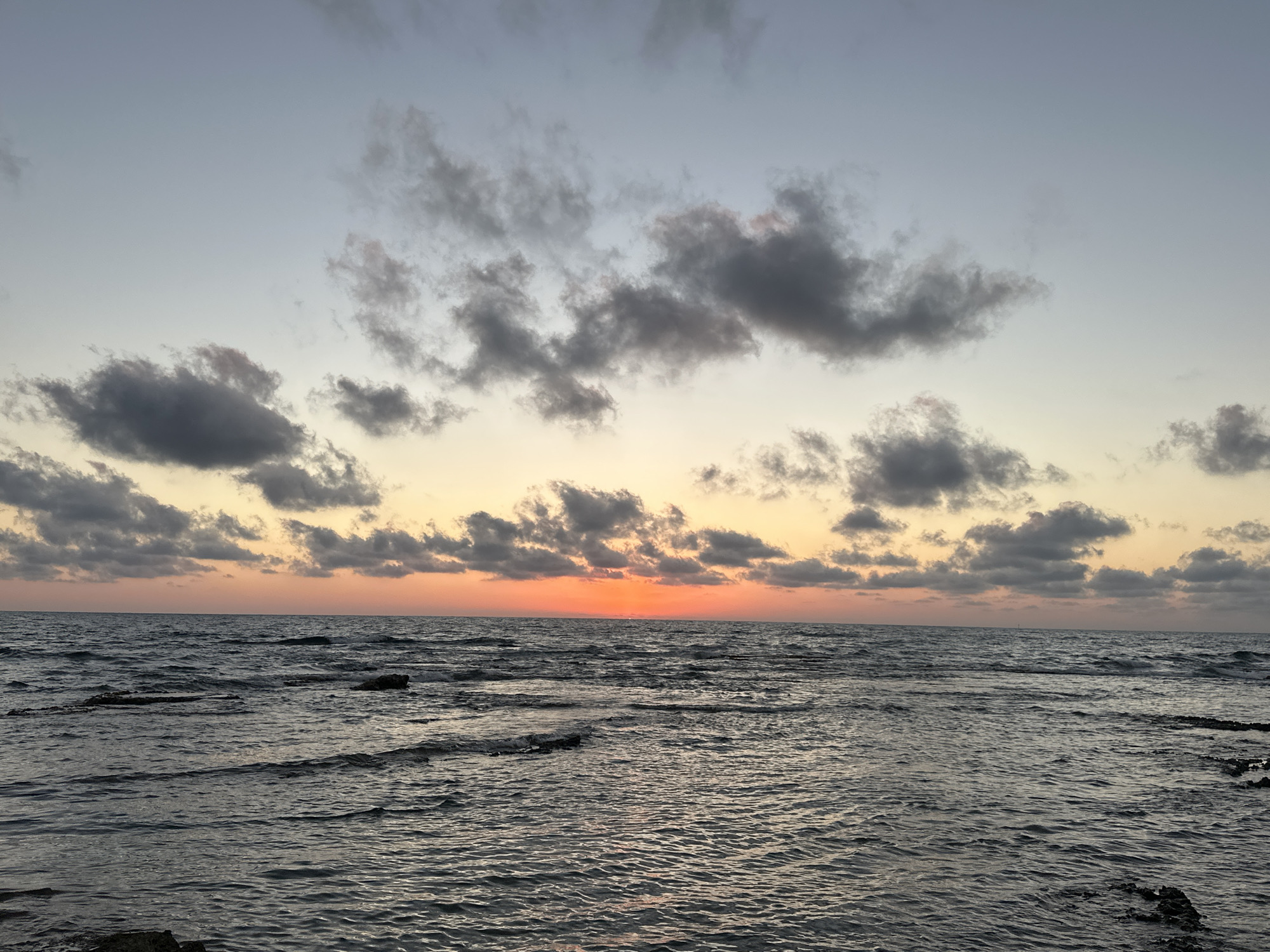 A serene sunset scene unfolds over the Mediterranean Sea near Haifa, Israel. The sky transitions from a pale blue at the top to warm hues of orange and yellow near the horizon, where the sun is setting. Scattered, dark clouds drift across the sky, some tinged with pink from the setting sun. The sea is choppy, with small waves lapping against rocky outcrops and the shore, visible in the foreground. The overall mood is peaceful and tranquil, characteristic of a coastal evening. No people or text are visible in the image.