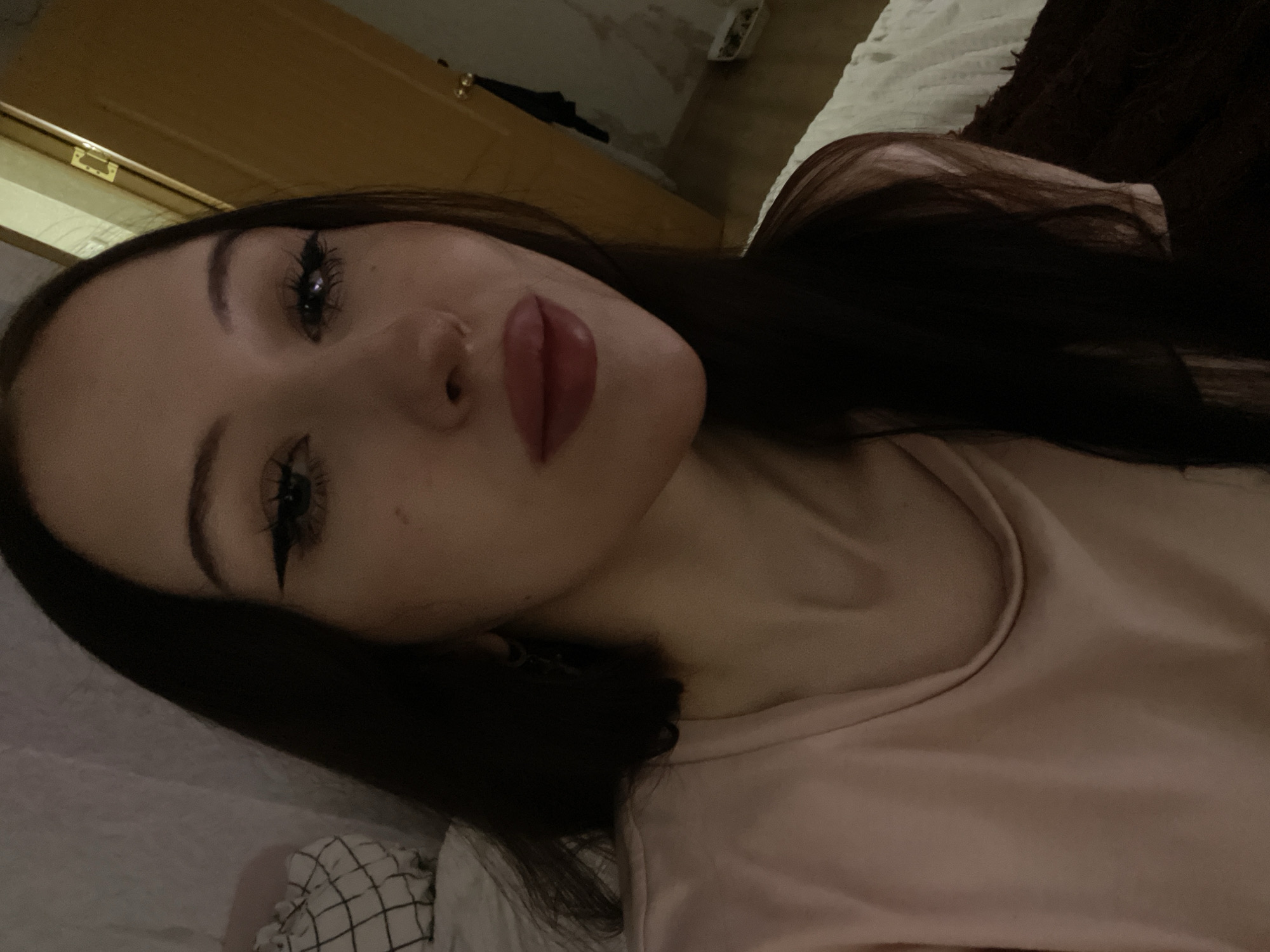 A young woman with long, dark hair and heavy eye makeup is taking a close-up selfie. She is wearing a light pink t-shirt. Her lips are full and appear to be wearing lipstick. The background is dimly lit, with a doorway visible on the right side of the image. There is a white textured surface behind her to the left, possibly a pillow or bedding. The image quality is low, with grain and limited detail. There is no indication of the specific location within Votkinsk, Russia.