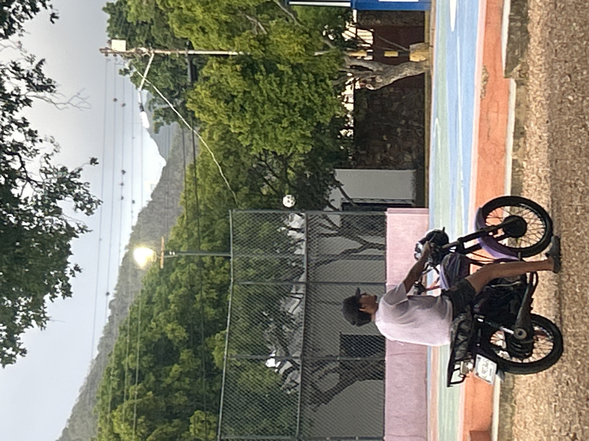 The image depicts a young man on a purple motorcycle, facing right, in what appears to be a public park or recreational area. The man is wearing a light-colored t-shirt, dark shorts, a black baseball cap, and dark shoes. The motorcycle is parked on a gravel surface. In the background, there is a fenced court, possibly a basketball or tennis court, with a colorful surface. Trees and hills are visible behind the court. A streetlamp is lit in the background, suggesting it might be dusk or overcast. The overall impression is that of a casual, everyday scene in a community setting. This location is identified as being in Santa Ana, Venezuela.