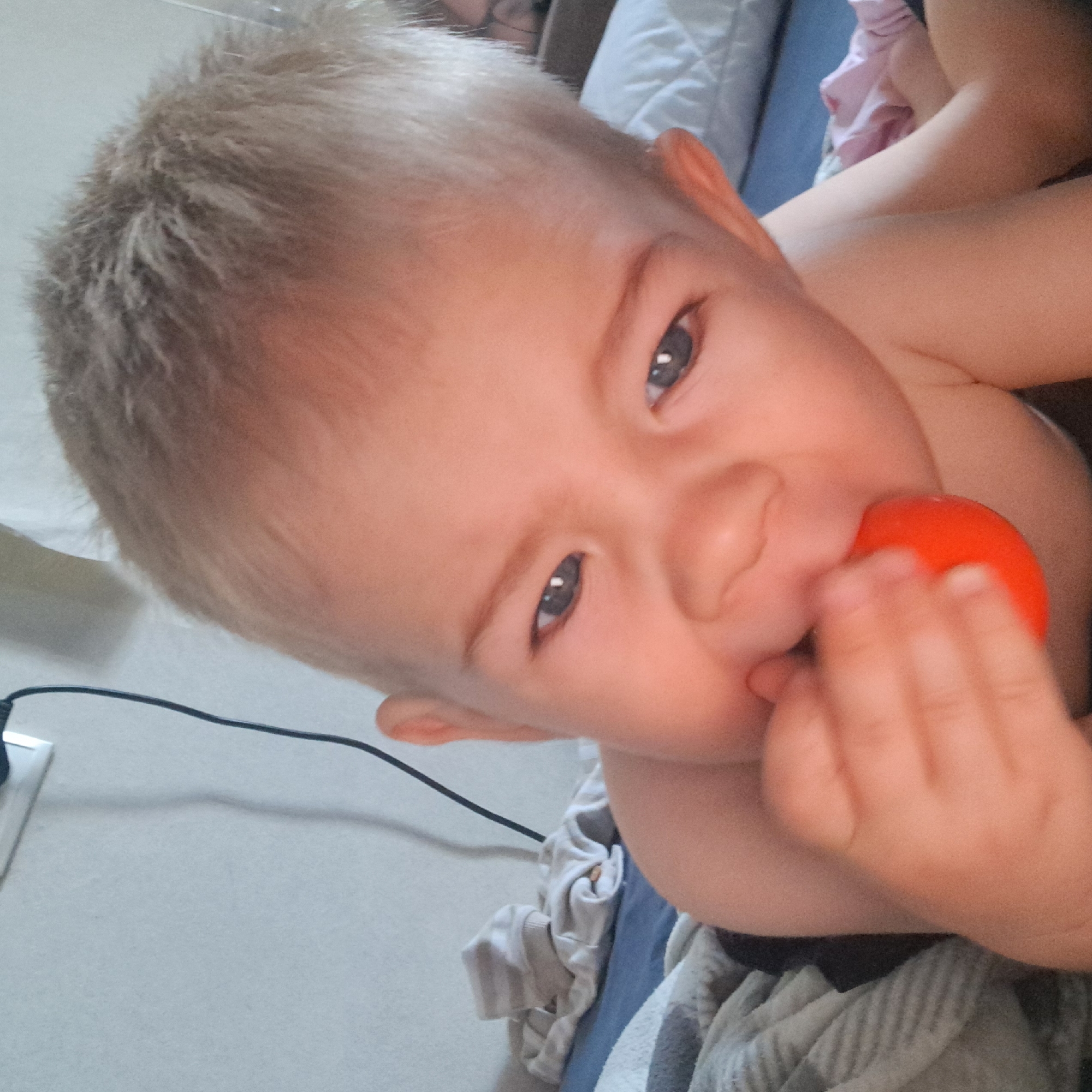 The image is a close-up, eye-level, indoor shot of a blonde-haired baby who is lying down and appears to be putting a small, round, orange object into their mouth. The baby is facing the camera and is looking slightly to the left. Their eyes are open, and their expression is somewhat concerned or perhaps slightly pained. The background is out of focus, but it appears to be a bed with bedding and some folded clothing. There is also a black cord and a white object on the wall in the upper left corner of the image, suggesting the presence of an electrical outlet and cord. The lighting is soft and warm. The scene is likely a domestic setting, such as a bedroom. There is no information in the image to suggest the location in the city of Krasnoyarsk, Russia.