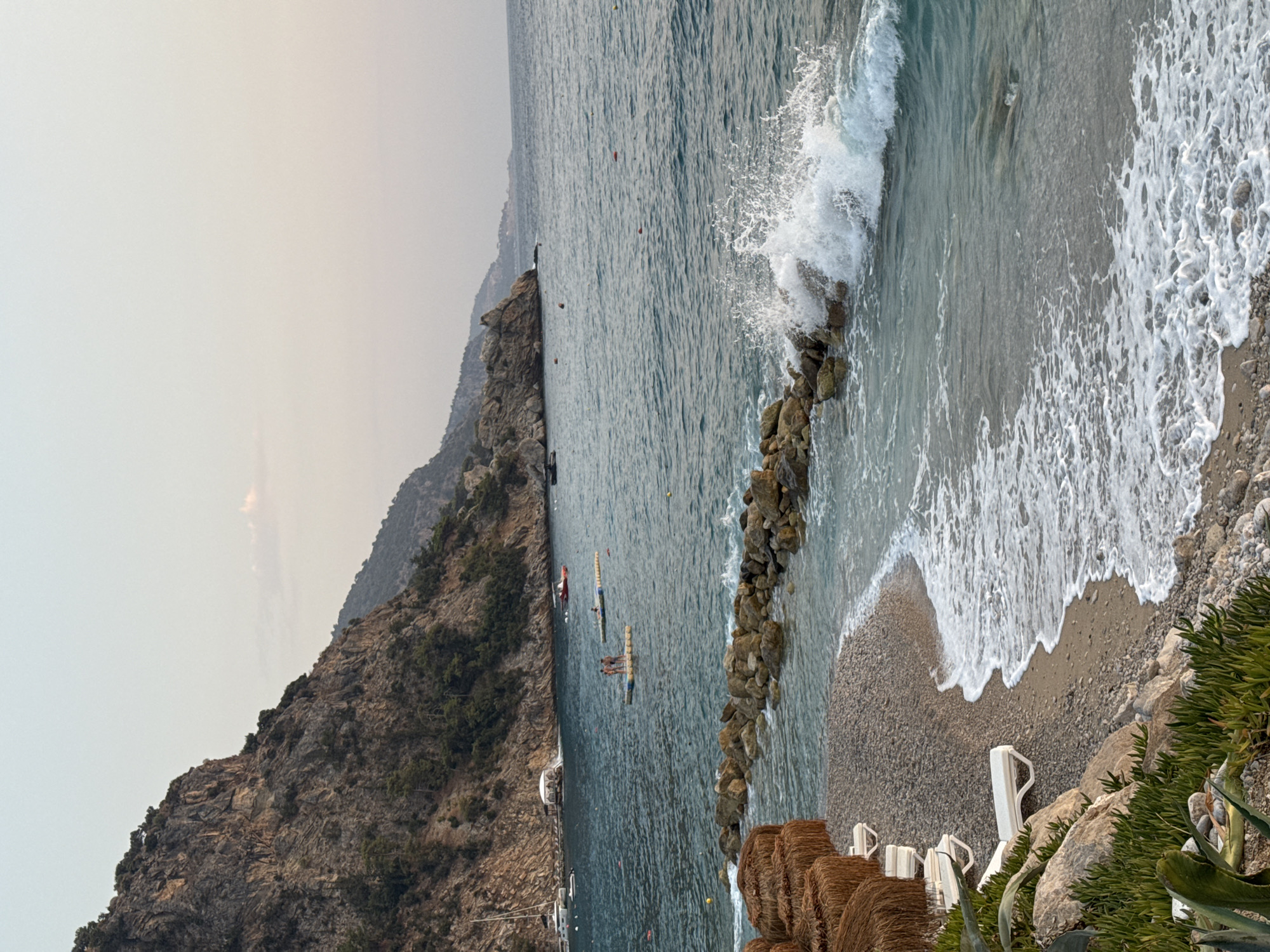 The image captures a serene coastal scene in Fethiye, Turkey. In the foreground, a pebble beach is visible, with several empty white lounge chairs and thatched umbrellas indicating a resort or beach club setting. Lush green vegetation and large rocks are also present on the beach. The sea is a deep blue, with gentle waves lapping at the shore. A breakwater made of large rocks juts out into the water, where a wave is crashing, creating a spray of white foam. In the middle ground, several people are seen standing on floating platforms in the water. The background features a dramatic, tree-covered rocky mountain range that slopes down to the sea. A small white building, possibly a restaurant or structure, is situated near the water's edge. The sky is a pale, hazy blue, suggesting it might be late afternoon or early morning. The overall impression is one of a tranquil holiday destination.