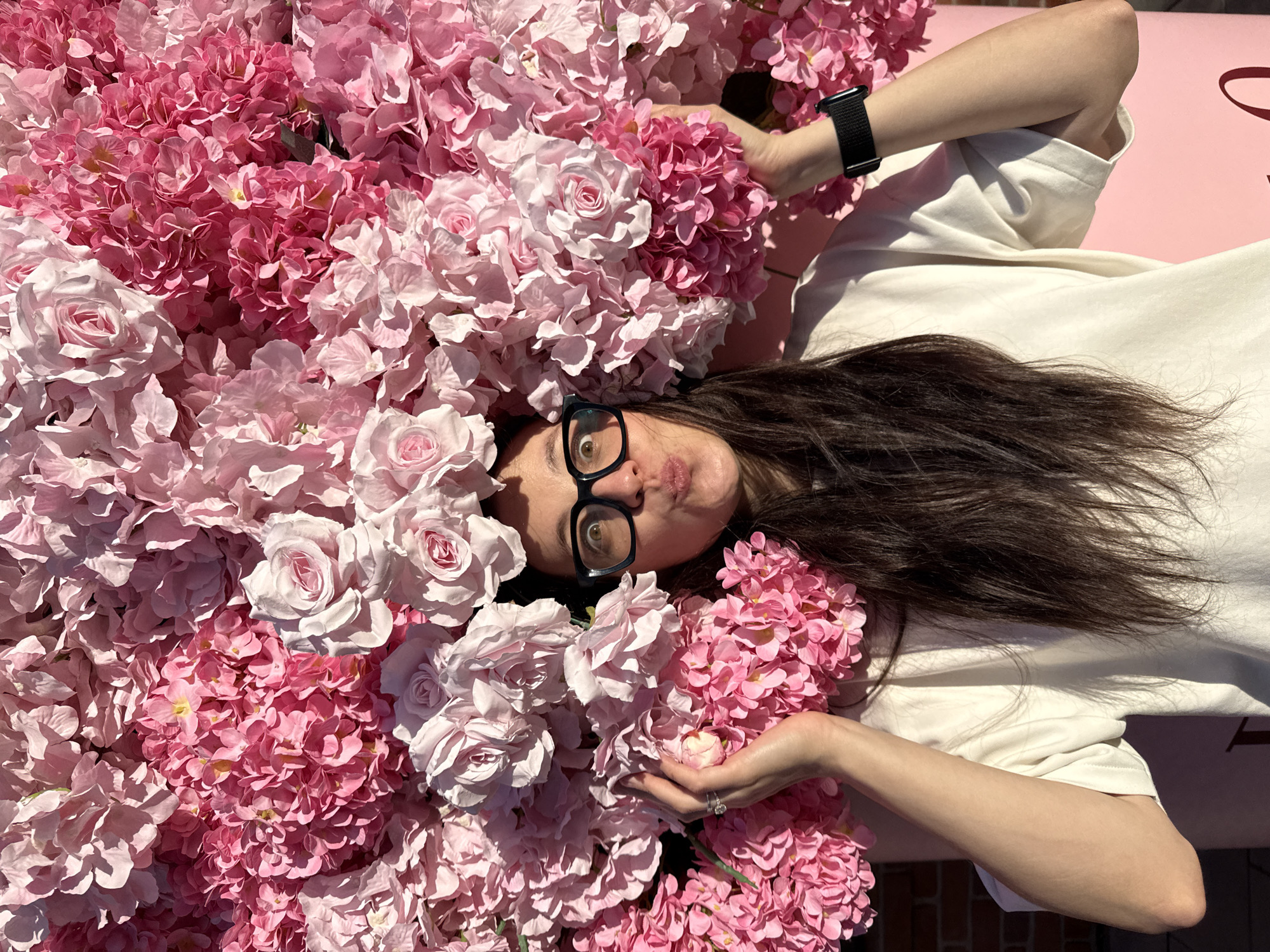 A woman with dark hair, wearing glasses and a white t-shirt, poses with a large floral arrangement of pink and light pink flowers, resembling roses and hydrangeas, which forms a backdrop and a sort of crown around her head. Her mouth is pursed in a playful expression. She is holding some of the flowers with her hands, one of which has a ring on her finger. A black smartwatch is visible on her left wrist. The background consists of more flowers and a hint of a brick wall and a pink structure with text that is mostly obscured. The scene appears to be outdoors, with sunlight casting shadows on the woman's arms and shirt. No specific location in Kazan, Russia, can be identified from the image.