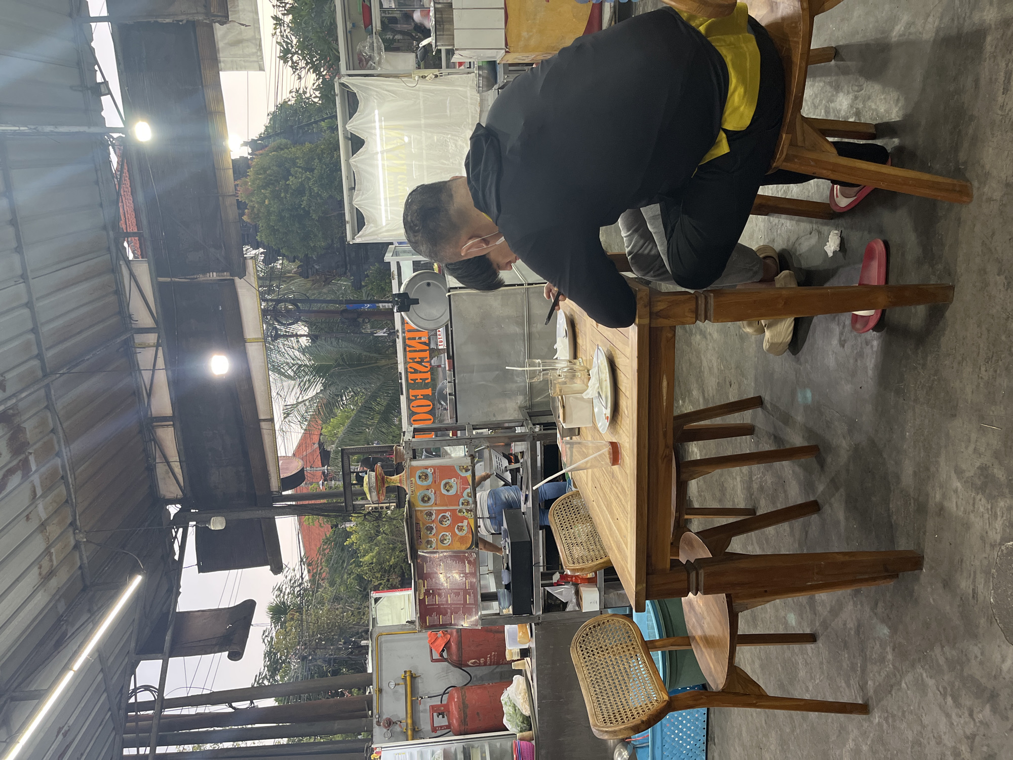 The image shows an outdoor dining area at a food stall in Kuta Selatan, Indonesia. Two individuals are seated at a wooden table. The person in the foreground, wearing a black hoodie and grey pants, is looking down at their phone. Another person, partially obscured, is seated opposite them. On the table are plates, glasses, and napkins, suggesting they are in the process of eating or have just finished. In the background, the food stall is visible with cooking equipment, gas cylinders, and menus displayed. The overall environment appears casual and functional, typical of a local eatery.