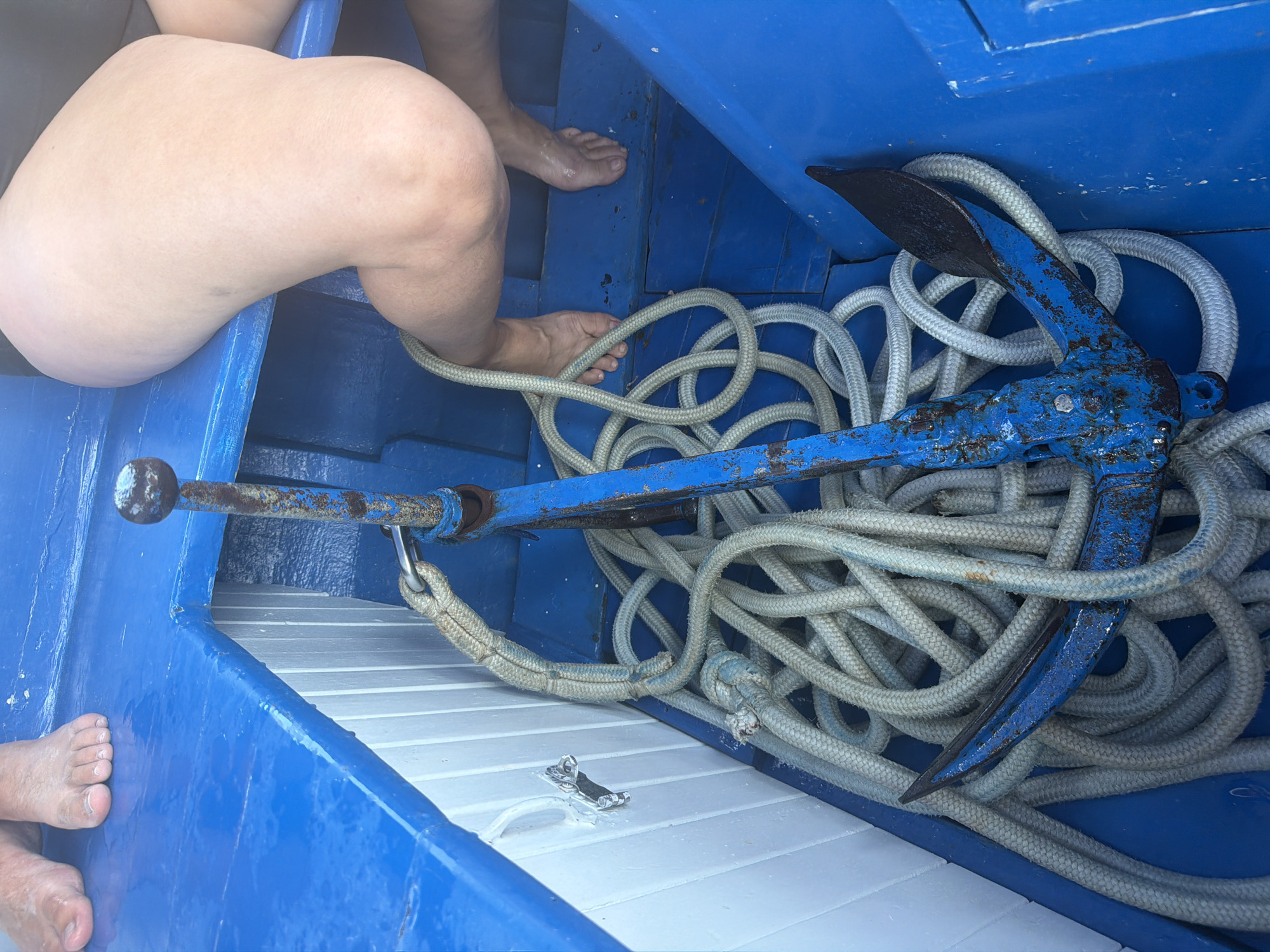 The image shows a blue wooden boat interior with a rusty blue anchor and coiled white rope in the foreground. In the background, the bare legs and feet of two people are visible, suggesting they are seated or resting on the boat. The environment appears to be outdoors, likely on the water, given the presence of the anchor and boat. The location is identified as Donji Štoj - Shtoji i Poshtëm, Montenegro.