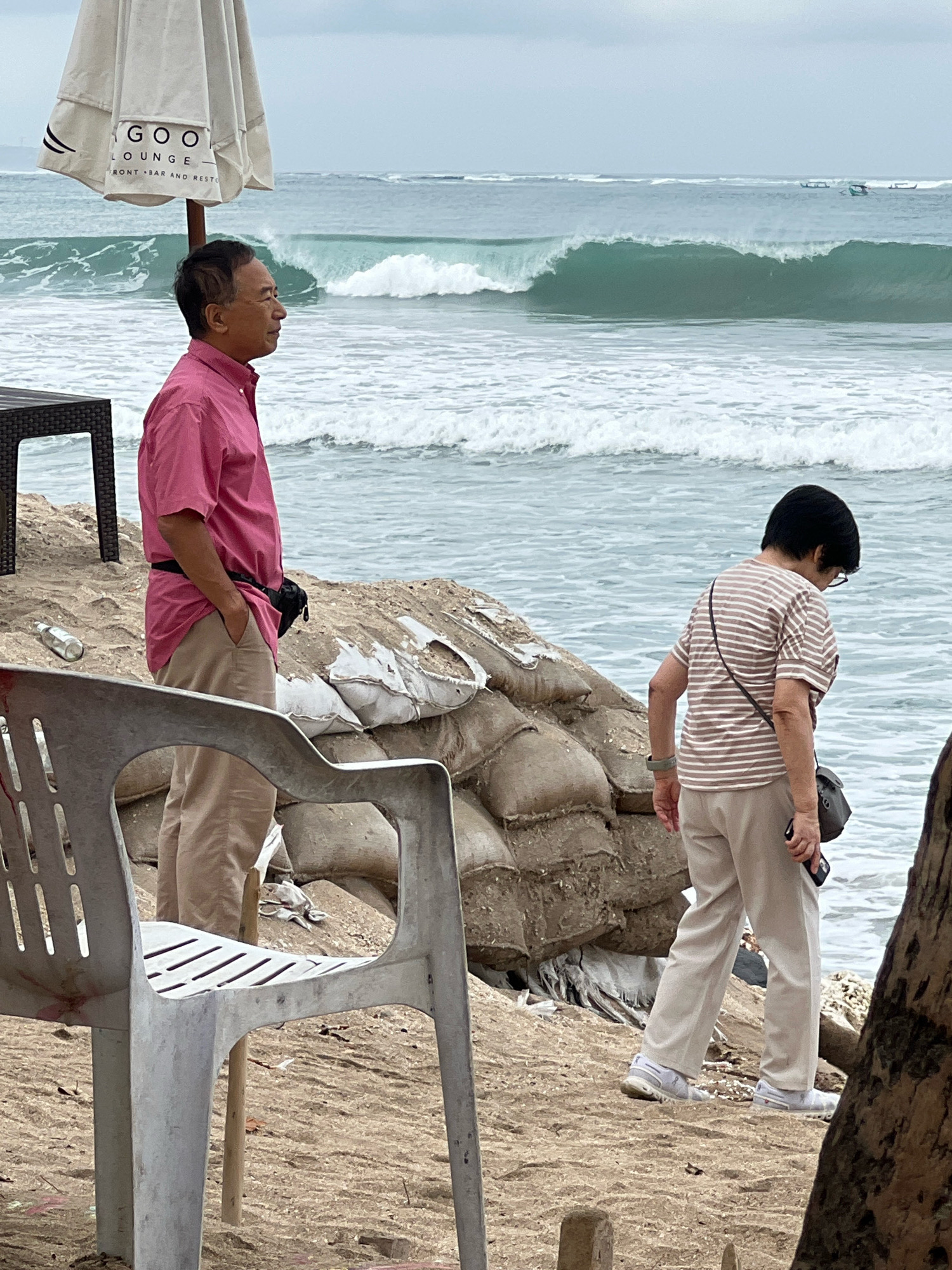 An outdoor, medium shot captures two people on a sandy beach in Kuta, Indonesia. In the foreground, a weathered white plastic chair is positioned on the sand. Behind it, a man, dressed in a pink collared shirt and tan pants, stands with his hands in his pockets, facing the ocean. He wears a black fanny pack around his waist. To his right, a woman in a horizontally striped t-shirt and tan pants walks away from the camera towards the ocean. She carries a small dark bag slung across her body and holds a phone in her right hand. The background reveals a tumultuous sea with a large wave breaking, characteristic of Kuta's surf. Sandbags are stacked to the right of the man, indicating some form of coastal defense. An umbrella with the text "GOO LOUNGE" is visible in the upper left corner, suggesting the presence of a beachfront establishment. The sky is overcast, contributing to a muted color palette.