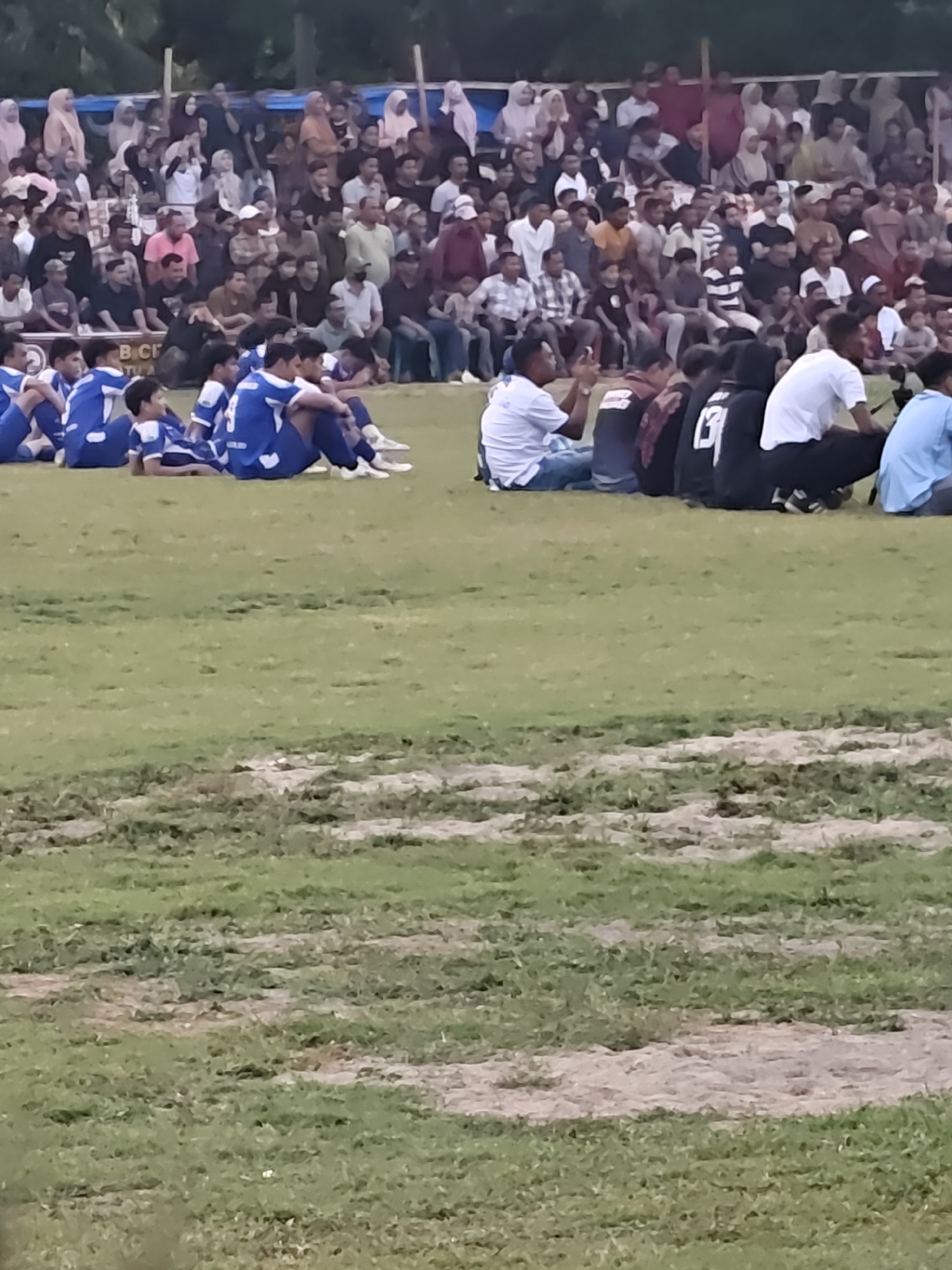 The image depicts an outdoor event, likely a sports match, with a crowd gathered in the background and a group of individuals seated in the foreground on a grassy field. The crowd in the background consists of many people, some standing and some sitting, under a blue and white tarp structure. They are dressed in various casual attire, with some women wearing headscarves. The seated individuals in the foreground appear to be a team, possibly soccer players, as many are wearing matching blue and white uniforms. Some are sitting cross-legged, while others are leaning back or lying down. The environment is a grassy field with some patches of bare soil. The overall scene suggests a community gathering or sporting event taking place in Desa Meunasah Blang, Indonesia.