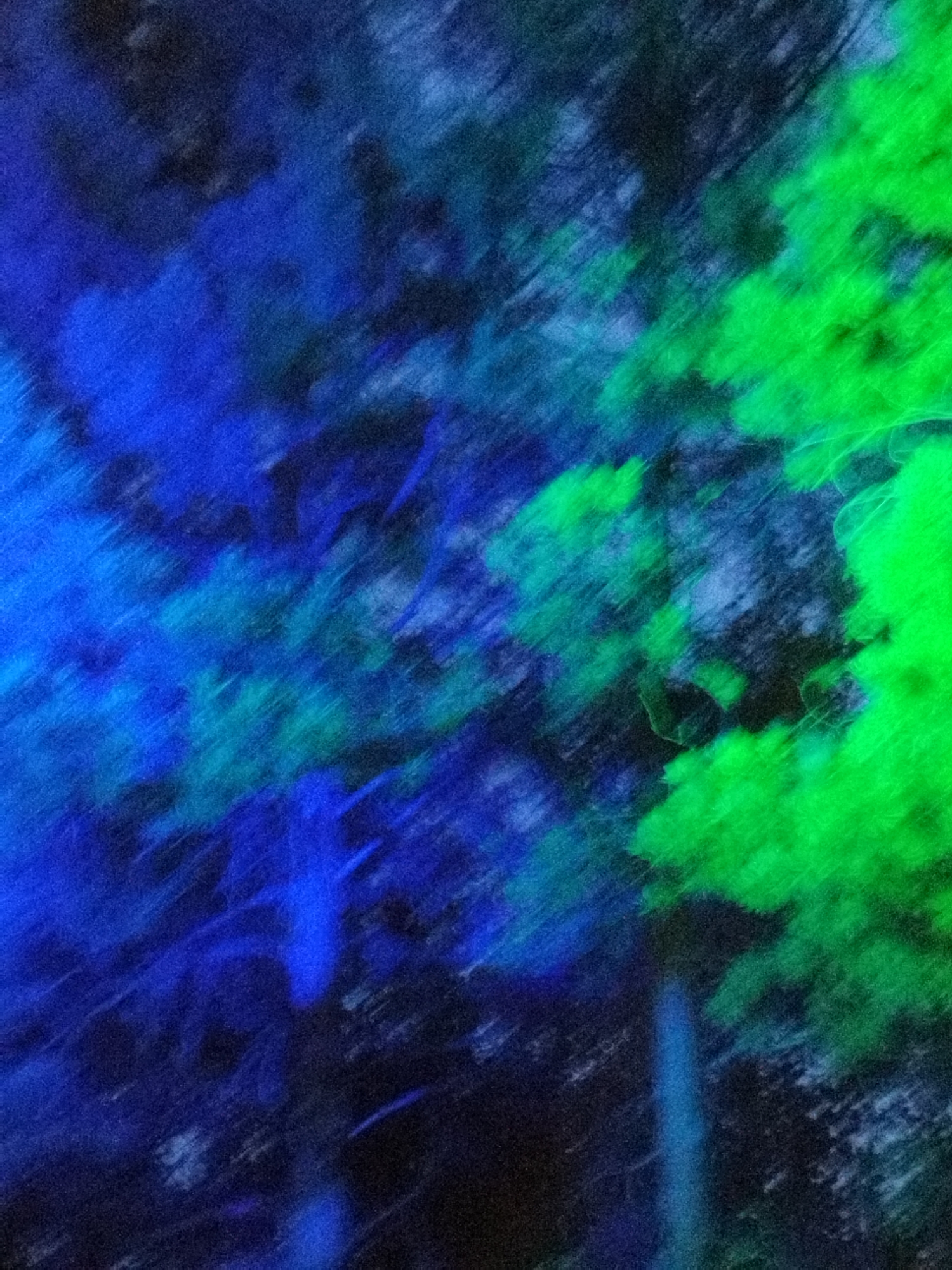 This is an abstract, impressionistic image with a blurred focus. It features diagonal strokes of vibrant blues and greens, set against a dark, possibly black, background. The interplay of light and shadow creates a sense of movement and depth. There are no identifiable people, subjects, or objects, and no discernible actions or interactions. The environment is not specified by any visual cues within the image itself. Therefore, without additional information, it is impossible to determine if this image is connected to the city of Surgut, Russia.
