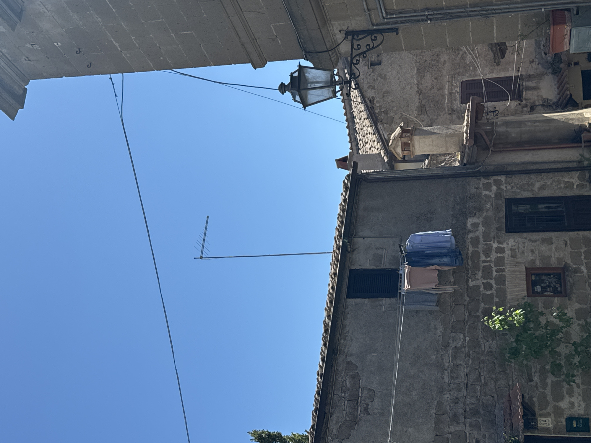 An upward-angled, exterior daytime shot captures a narrow street scene in Castelnuovo di Porto, Italy. On the right, the facade of an old stone building dominates, featuring small, shuttered windows, exposed brick, and a framed picture. Laundry hangs on a rack near the building's side. A portion of the blue sky is visible, with a television antenna extending towards the left. Overhead, the edge of a building is partially visible, obscuring more of the sky. A black, decorative street lamp is mounted on the building on the right. Wires connect the lamp to the building and run across the open space.