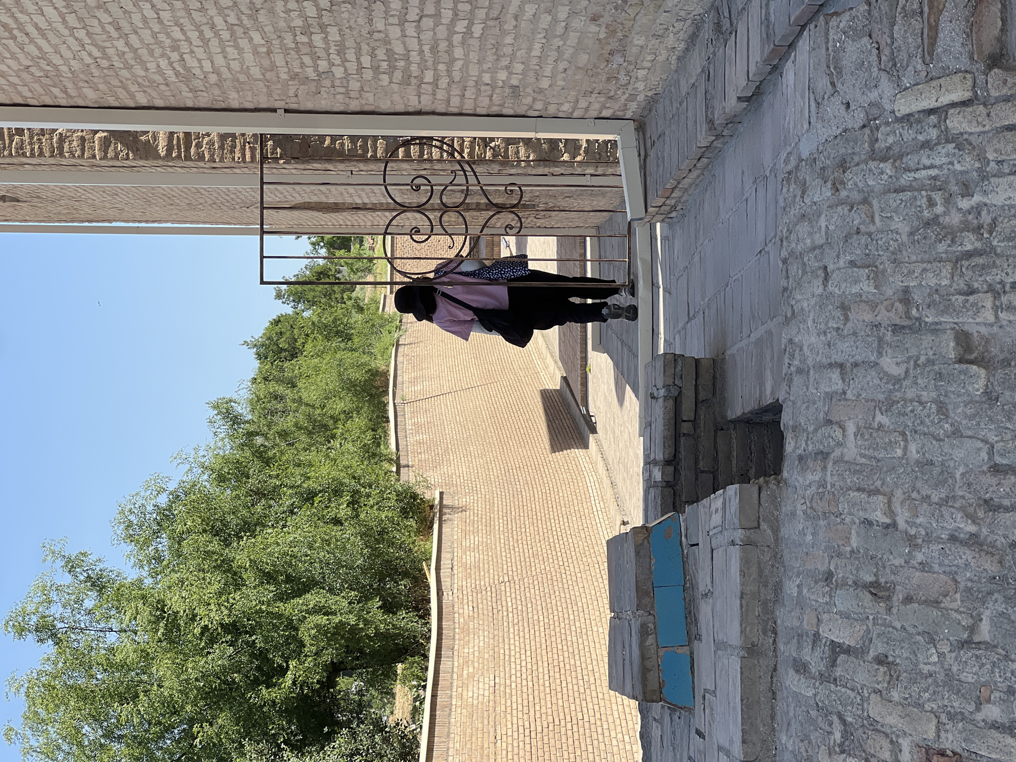 A person in a black hat, pink shirt, and black pants walks away from the camera through a decorative metal gate towards a brick building in Samarkand, Uzbekistan. The person is carrying two bags, one black and one patterned. The ground is paved with bricks. To the left, a brick wall runs along a pathway lined with trees. The sky is clear and blue.