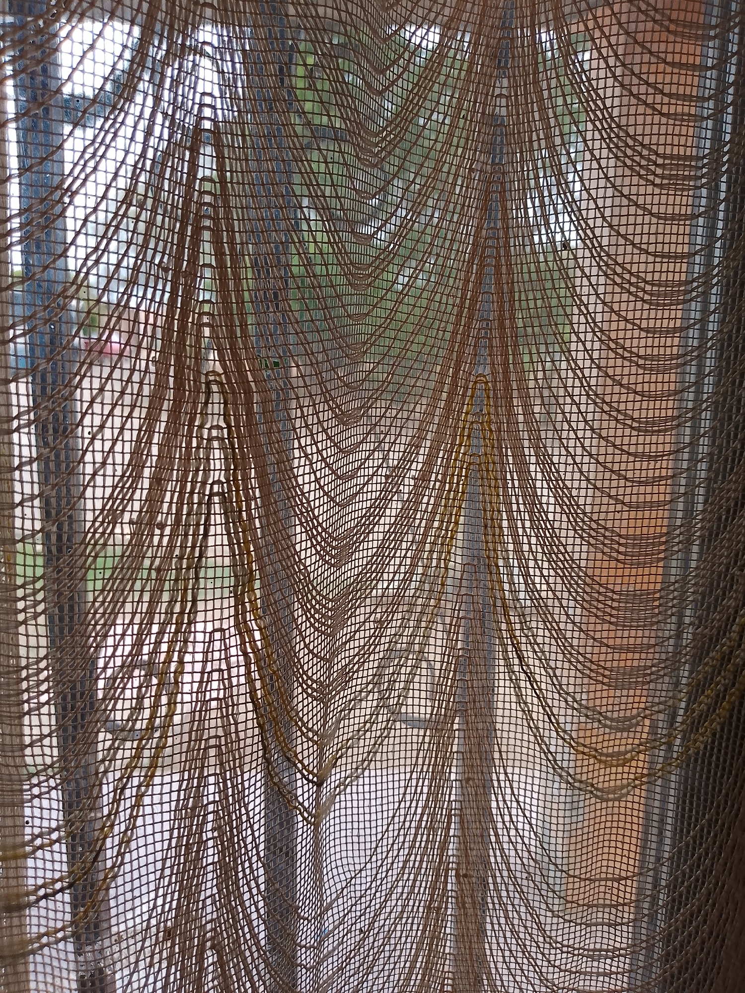 This image is a close-up, low-angle shot of a textured, sheer fabric curtain hanging in front of a window, possibly in a home. The fabric is a light brown or beige color and has a loosely woven, net-like texture with horizontal threads creating a grid pattern and vertical threads that are thicker and more spaced out, forming draped, wavy lines. Some of these vertical threads appear to be frayed or have added yellow strands woven into them, creating a distressed or decorative effect.
Through the sheer fabric, the view outside is blurred and out of focus. It appears to be daytime, with natural light filtering through. Hints of greenery suggest trees or other foliage. In the background, there are also suggestions of buildings with some reddish-brown tones and possibly window frames. The overall impression is one of a casual, indoor setting looking out onto an outdoor environment.
The image does not contain any people or discernible actions. The focus is entirely on the texture and draping of the fabric and the diffused view beyond.
Regarding the location of Santa Ana, Venezuela, this image provides no specific visual cues to confirm or deny this location. The elements visible are generic enough to be found in many places. Without further information or distinguishing features within the frame, it is impossible to pinpoint the location as Santa Ana, Venezuela.