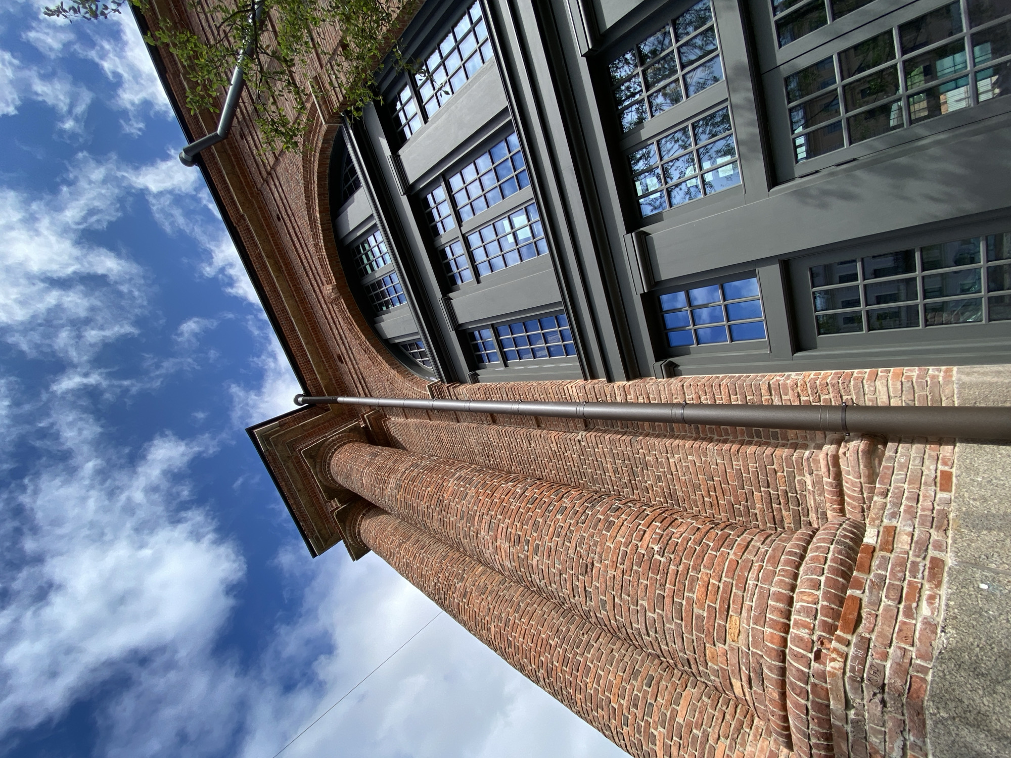 This is a low-angle, close-up exterior shot of a building in Saint Petersburg, Russia. The building features a red brick facade with a large arched window framed by dark grey trim. Several rows of multi-paned windows are visible. To the left, a section of the building shows two large, curved brick columns. A dark downspout runs vertically along the brickwork. The sky above is a vibrant blue with scattered white clouds. A few green tree branches are visible on the right side of the frame, suggesting the building is surrounded by greenery. The overall impression is of an old, possibly industrial or institutional, building with a blend of historical and modern architectural elements.
