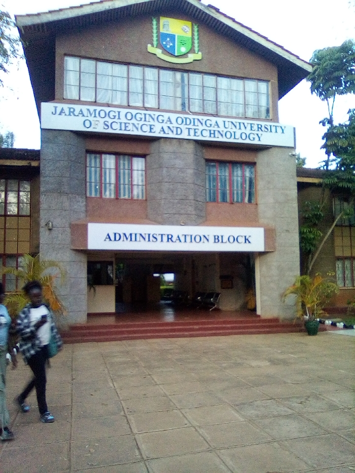 This image depicts the exterior of the Administration Block at Jaramogi Oginga Odinga University of Science and Technology, located in Bondo, Kenya. The scene is observed from a paved courtyard area in front of the building under what appears to be an overcast or diffused daylight sky.
The central structure is a multi-story building featuring a brown, stucco-like facade interspersed with grey concrete or stone cladding sections. A brown, sloped roof tops the building. A prominent white banner stretched across the upper level displays "JARAMOGI OGINGA ODINGA UNIVERSITY OF SCIENCE AND TECHNOLOGY" in blue capital letters. Below this, a second white banner positioned above the main entrance reads "ADMINISTRATION BLOCK," also in blue capital letters. Above the upper banner, near the roofline, a colorful university crest or emblem featuring a shield and foliage is affixed to the facade.
The building incorporates numerous rectangular windows with dark frames, some exhibiting red or maroon vertical elements internally, and white curtains or blinds are visible behind some panes. The main entrance is a recessed archway accessible via reddish-brown steps. Inside the entrance, a counter is visible to the left, and a row of dark chairs, possibly for waiting, is positioned to the right. Potted plants flank the entrance, and a security camera or light fixture is mounted on a grey column to the right of the upper signage.
In the lower-left foreground, two individuals are walking across the grey square-tiled paved surface, moving away from the camera towards the left. The individual closer to the camera is a young female with dark hair, wearing a dark plaid shirt over a light-colored top, dark pants, and light-colored shoes, carrying a dark shoulder bag. She is captured in mid-stride. A second individual is partially visible to her left, wearing light-colored pants and dark shoes, walking alongside her. The surrounding environment includes green trees and foliage on both sides of the building and in the background.