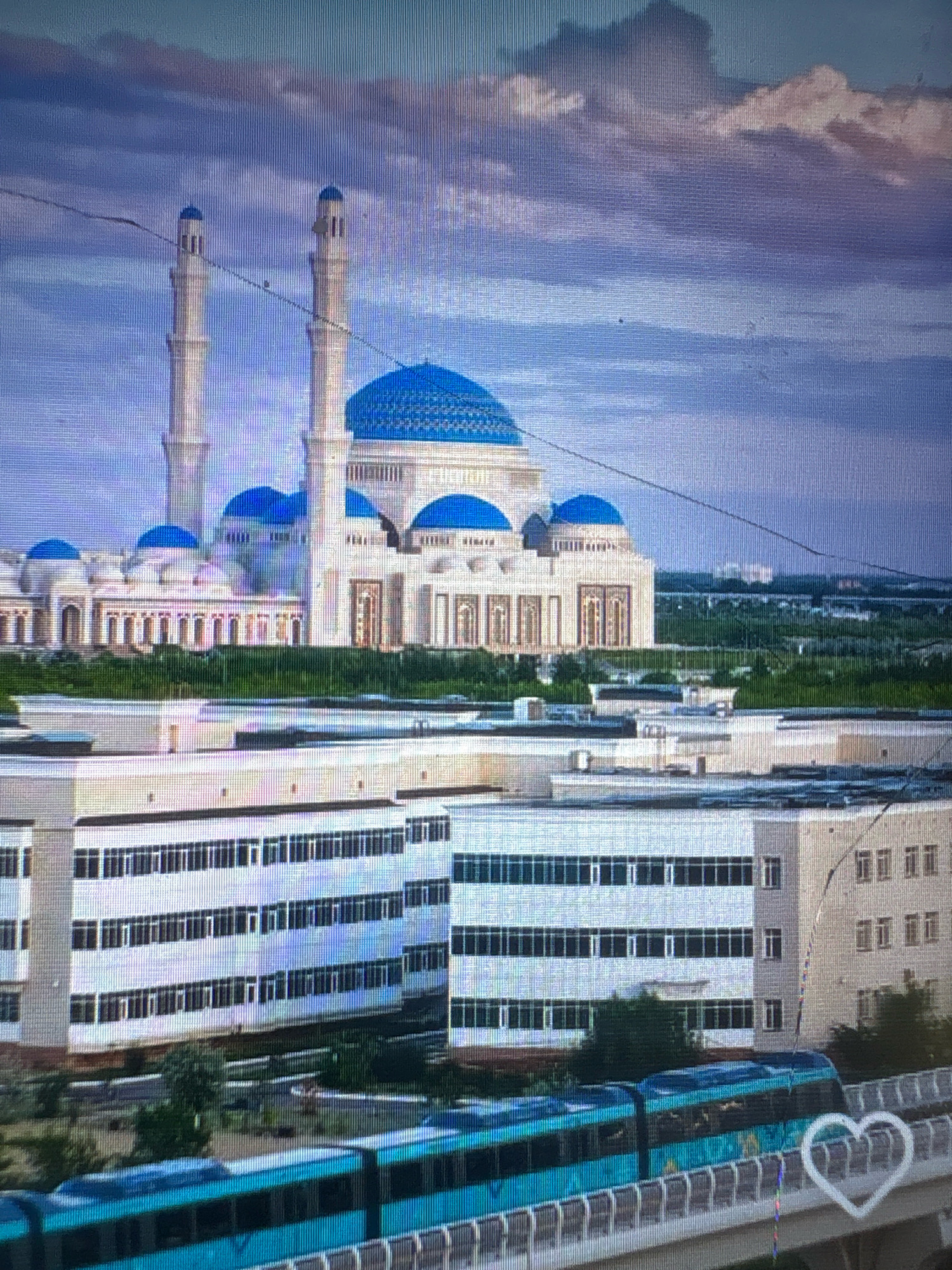 The image depicts the Palace of Peace and Reconciliation, a pyramidal structure with a large blue dome, situated in Astana, Kazakhstan. Two tall minarets, characteristic of Islamic architecture, flank the main building. In the foreground, a modern blue train travels on an elevated track, passing by low-rise buildings with rows of windows. The sky above is a mix of blue and dramatic clouds, suggesting a partly cloudy day. The surrounding area appears to be a blend of urban development and green spaces.