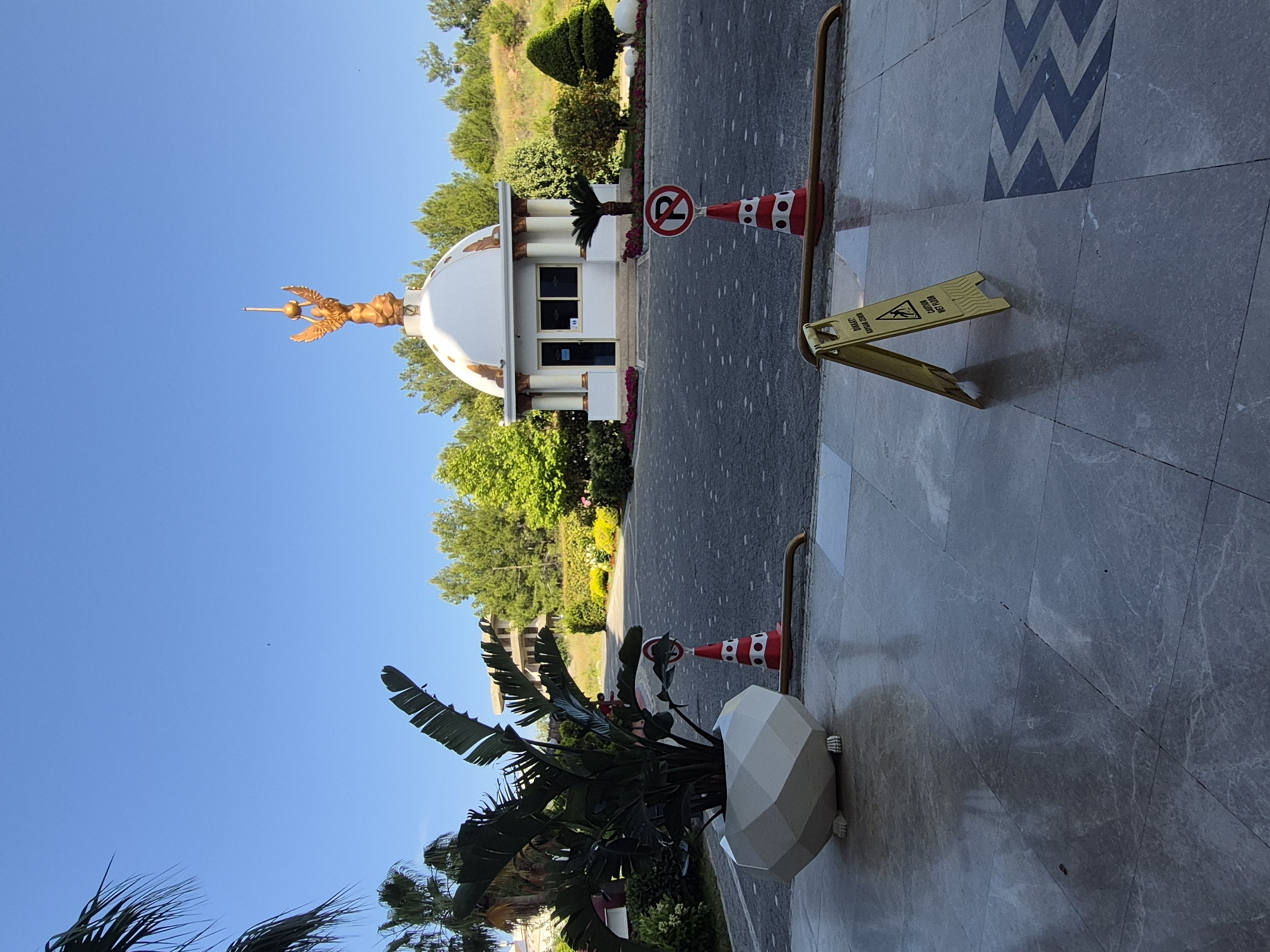 The photograph depicts an exterior scene in Alanya, Turkey. A small, white guardhouse with a dome topped by a golden angel statue is visible in the middle ground, adjacent to a paved road. A red-and-white striped traffic cone with a no-parking sign rests near the guardhouse. In the foreground, a grey marble-tiled surface with a blue geometric pattern is present. A yellow "caution wet floor" sign is also in the foreground, along with a large decorative flowerpot containing tropical plants. The sky is clear blue. Vegetation, including trees and shrubs, forms a backdrop behind the guardhouse.